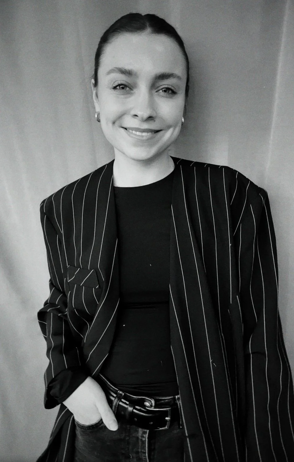 Schwarz-weiß-Foto einer lächelnden Frau mit dunklem Haar, die eine schwarze, gestreifte Jacke trägt und die Hand in die Tasche steckt.