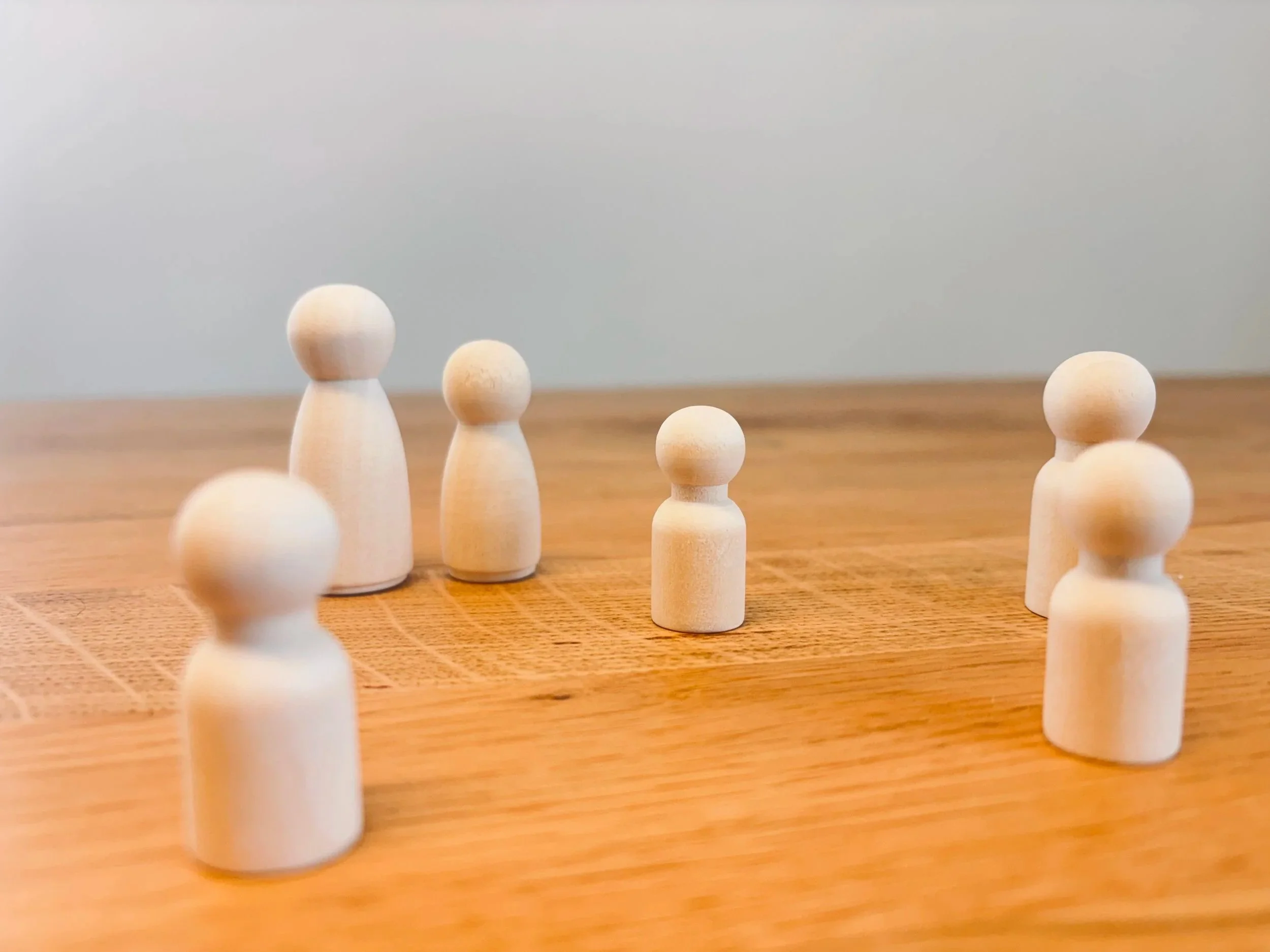 Mehrere kleine, helle Holzfiguren mit runden Köpfen und ovalen Körpern stehen auf einem Holztisch vor einem grauen Hintergrund.
