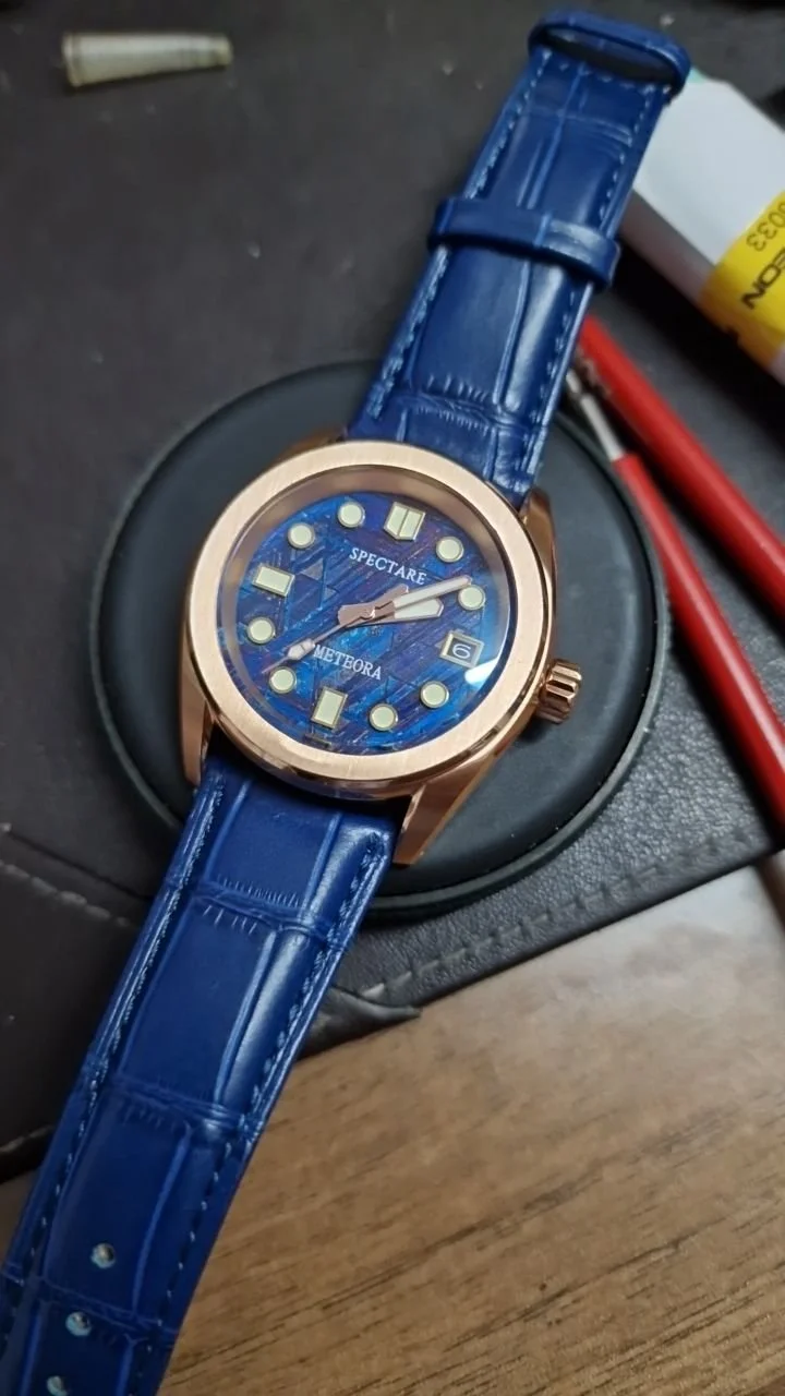 A wristwatch with a blue face and blue strap placed on a black surface, surrounded by tools and objects.
