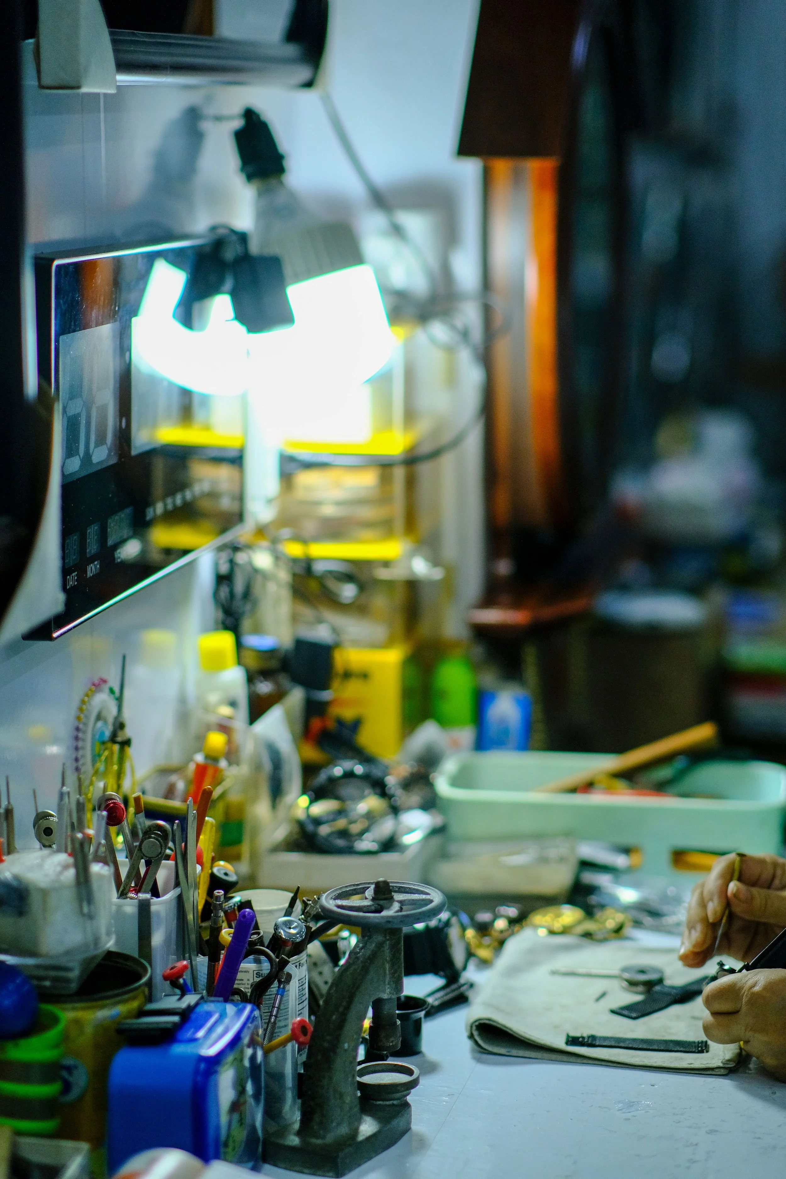 A cluttered workshop or repair station with various tools and equipment on the workbench, including pliers, screwdrivers, and a soldering iron, with a worker's hands holding a small object over a cloth.