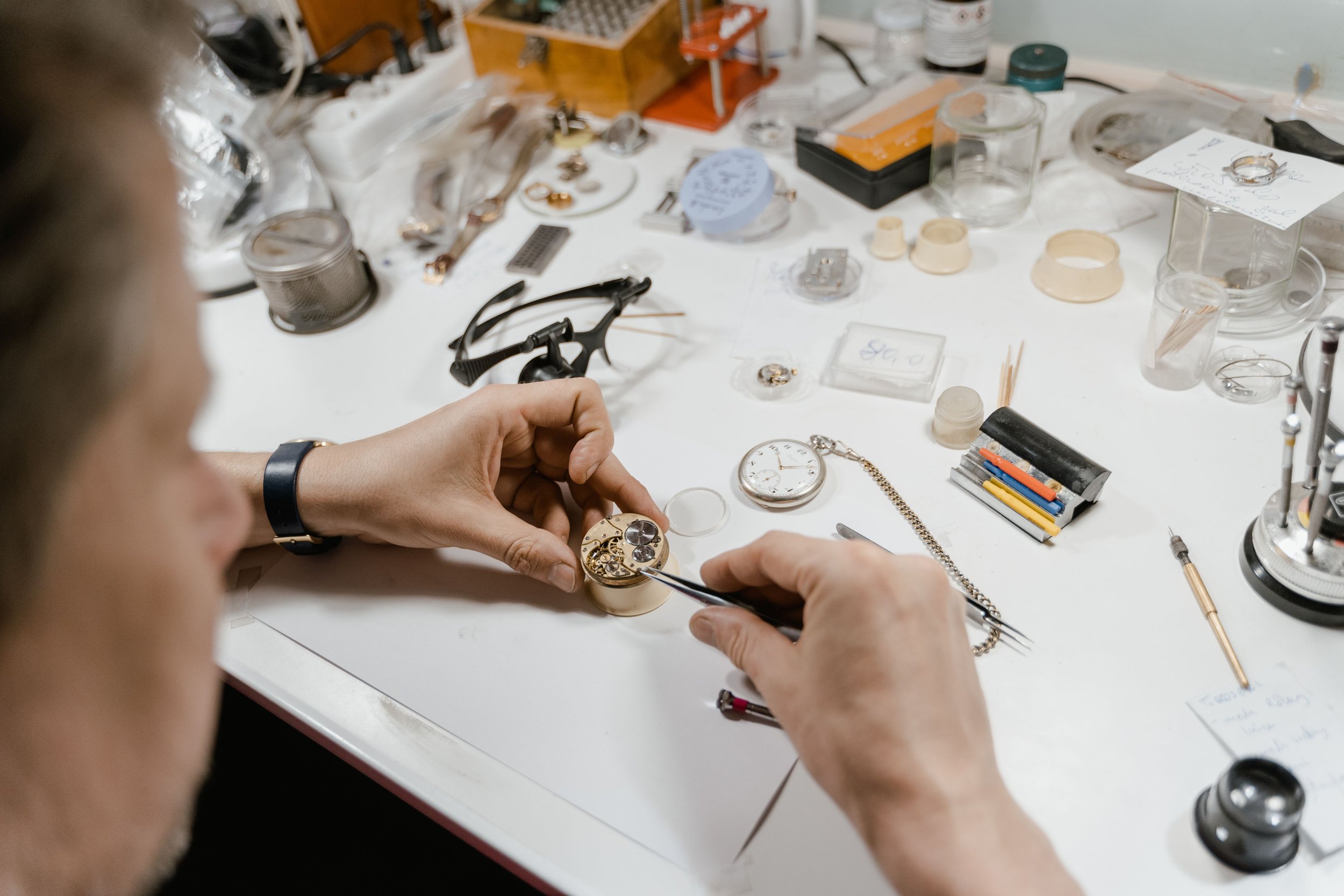Person working on repairing a wristwatch at a cluttered workbench with various tools and watch components.