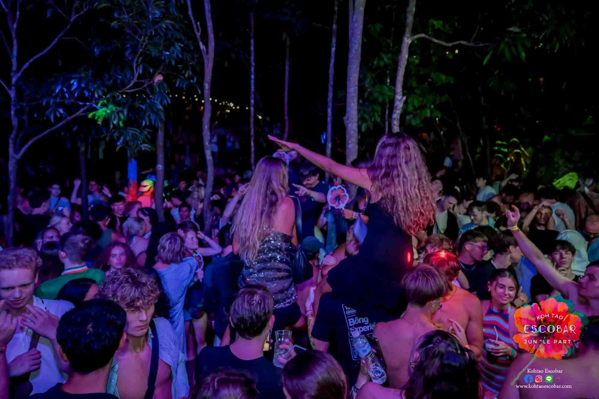 Crowd of people at an outdoor party or festival at night, with two women with long hair in the foreground, one pointing into the air. Colorful lights and a foliage backdrop are visible.