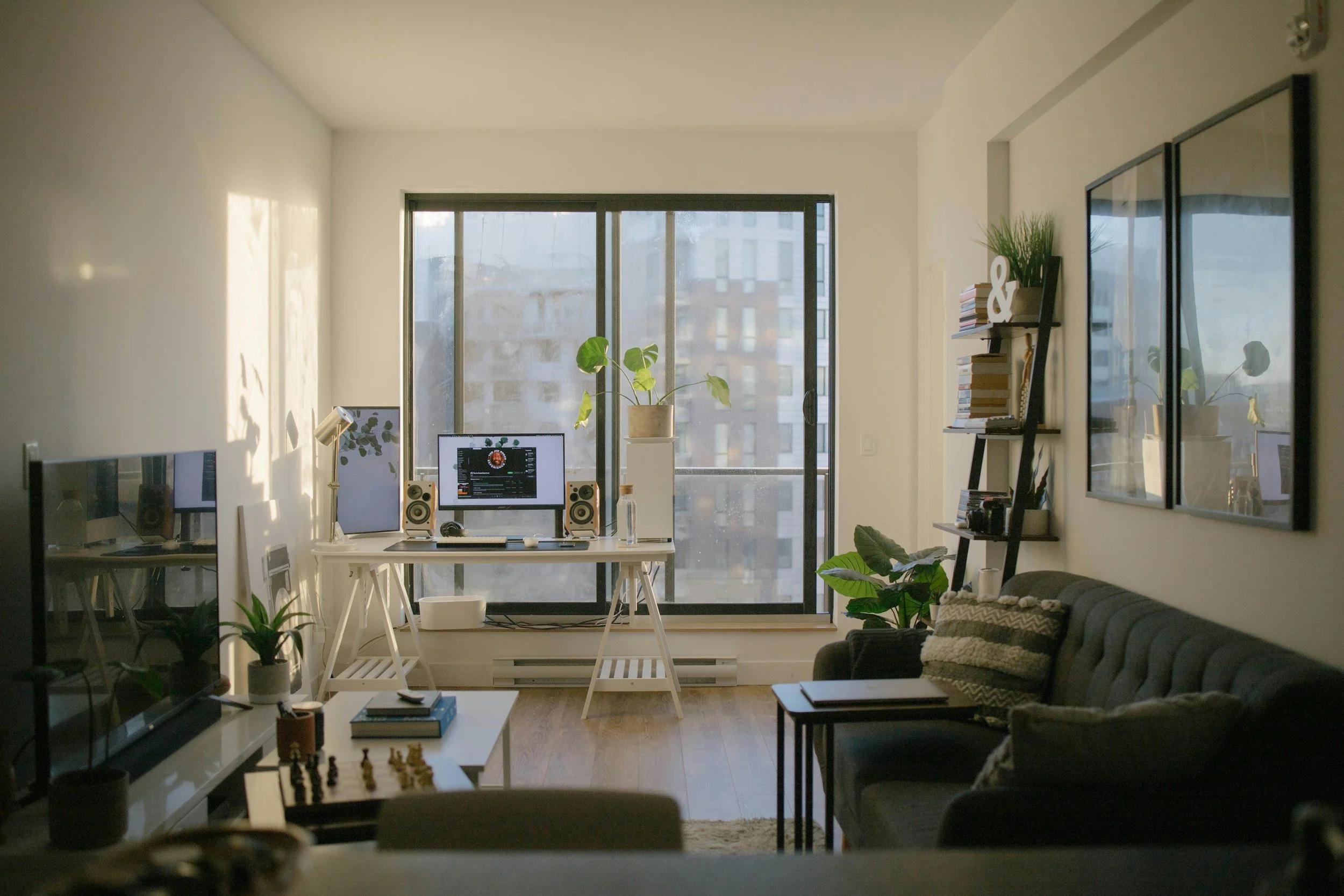 A modern apartment living room with a workspace by the window, potted plants, books, framed pictures, and a gray sofa with patterned pillows.