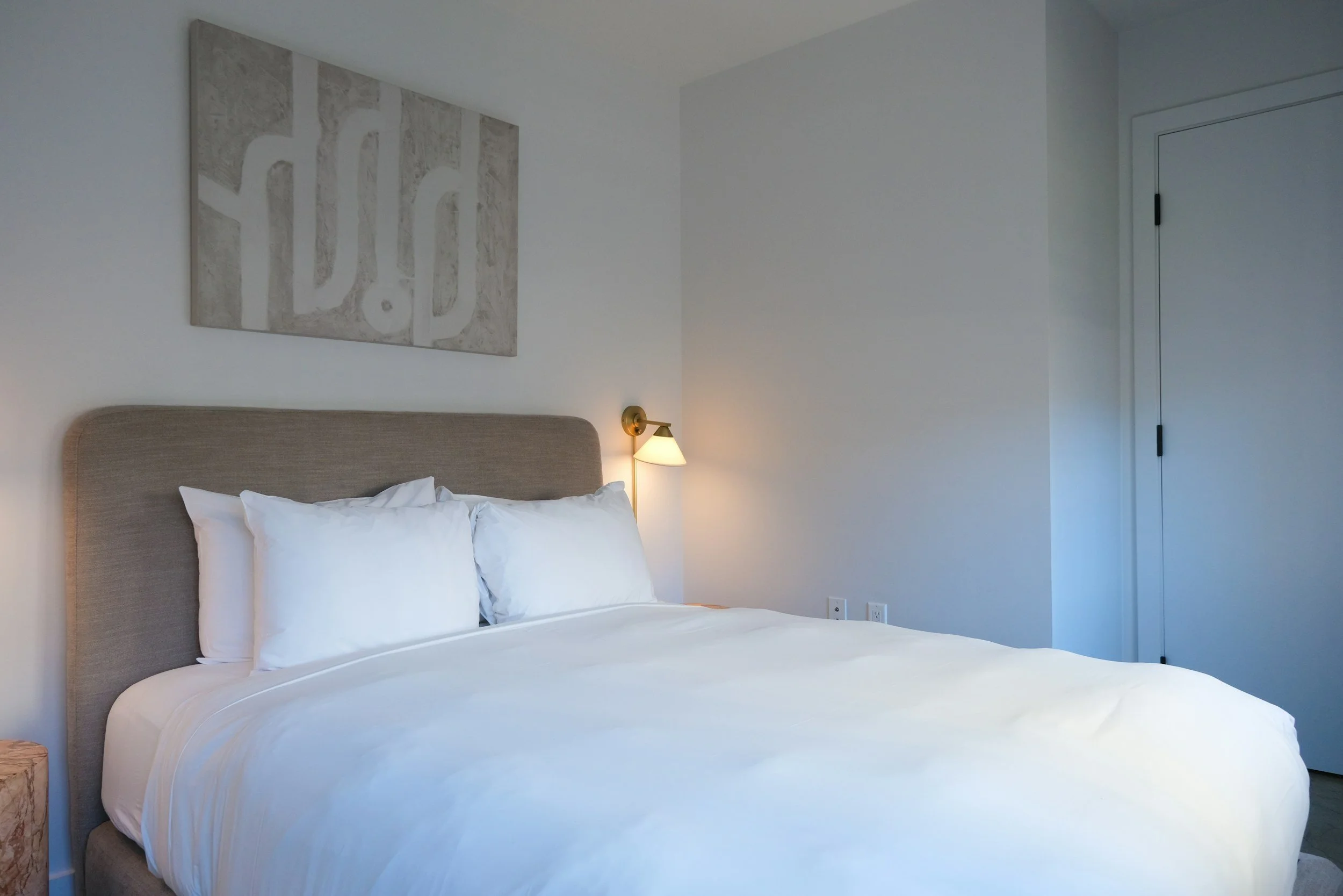 A neatly made bed with white linens, two pillows, a beige upholstered headboard, a wall-mounted wall lamp, a piece of abstract art on the wall, and a white door in the background.