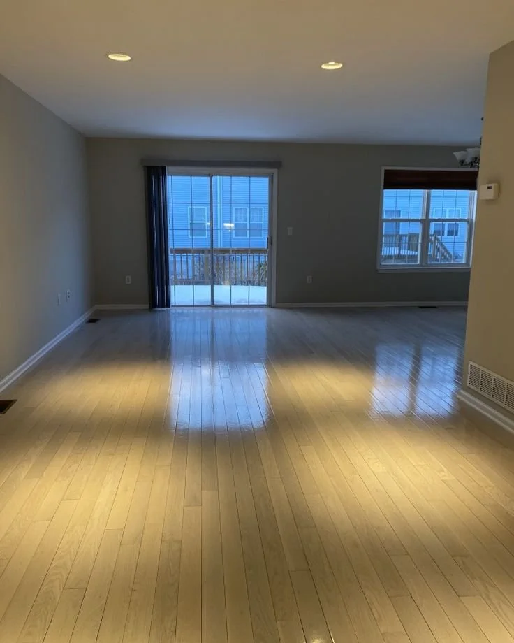Empty living room with hardwood floors, sliding glass door leading to a balcony, and window with blinds, in a modern apartment.
