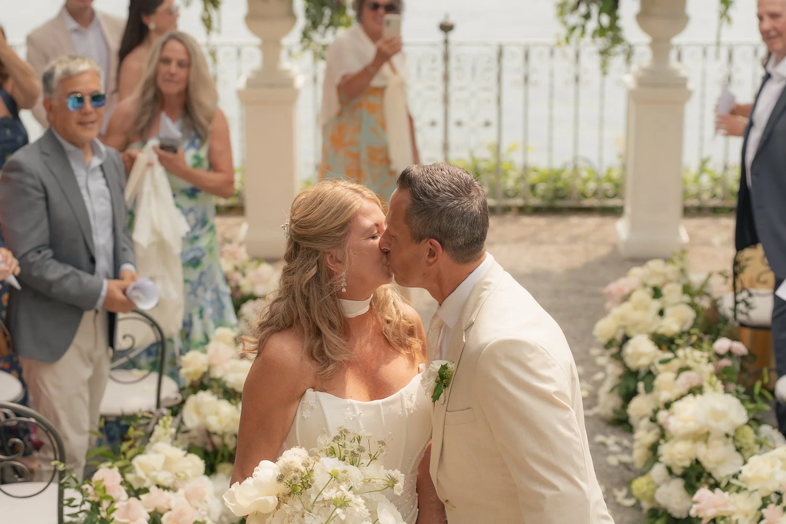 Una coppia di sposi si bacia durante il matrimonio, circondata da amici e parenti, con decorazioni floreali bianche e rosa.