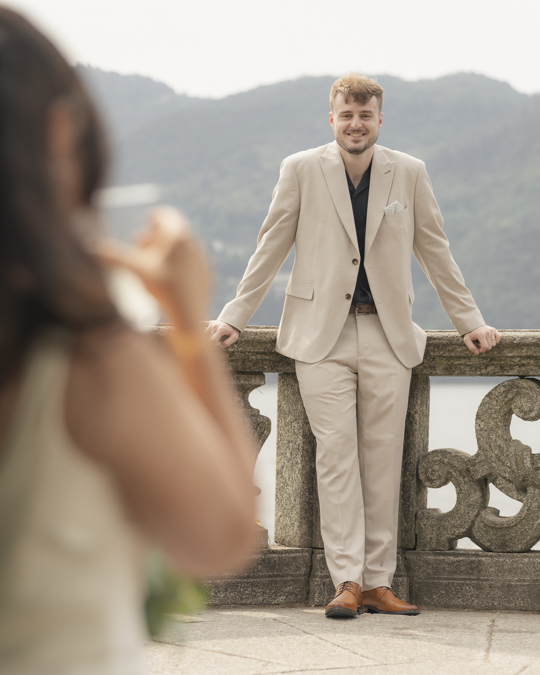 Uomo sorridente in abito beige che si appoggia a un muro di pietra con sfondo montano, mentre una donna in primo piano scatta una foto con il cellulare.