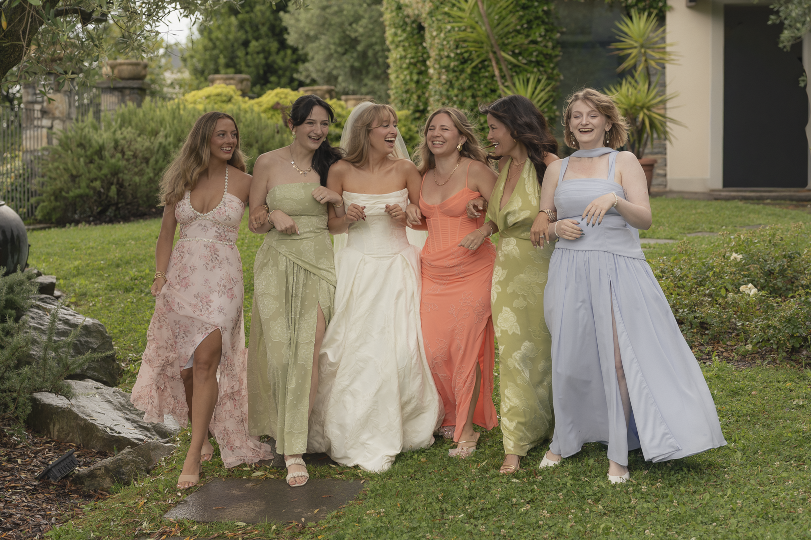 Sette donne sorridenti che camminano insieme in un giardino, indossando abiti da cerimonia colorati.