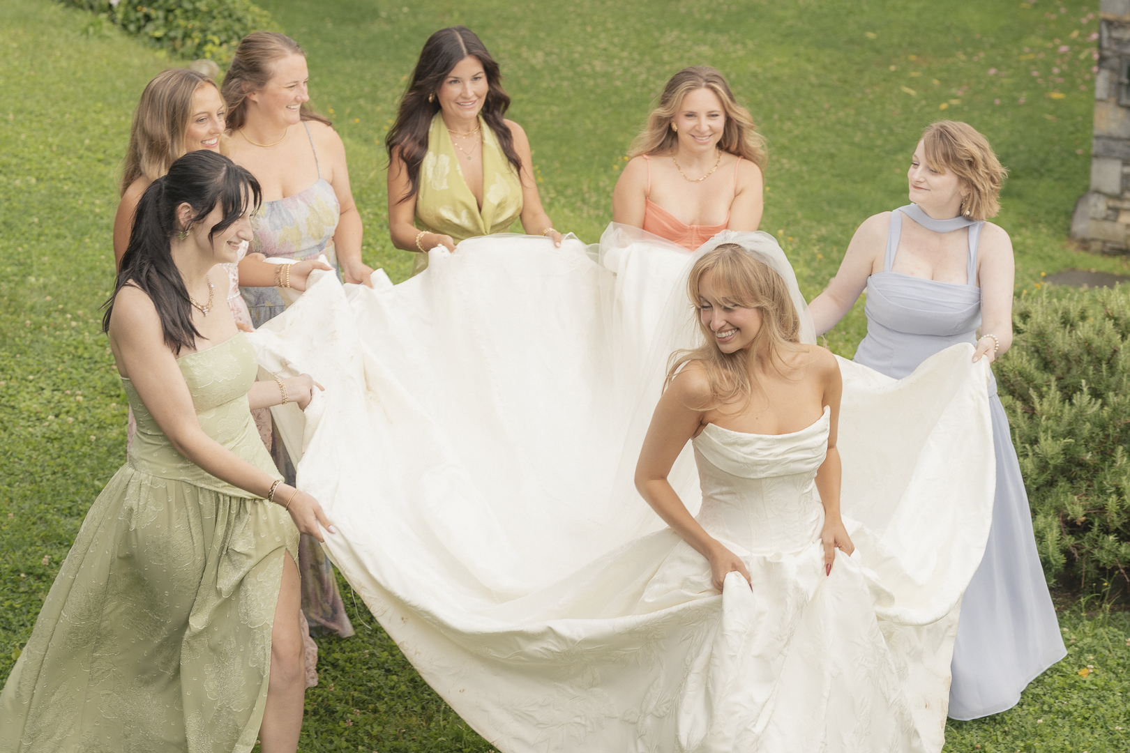 Un gruppo di donne felici in abiti eleganti, che sorridono e tengono una lunga stoffa bianca su un prato verde, in un giardino all'aperto.
