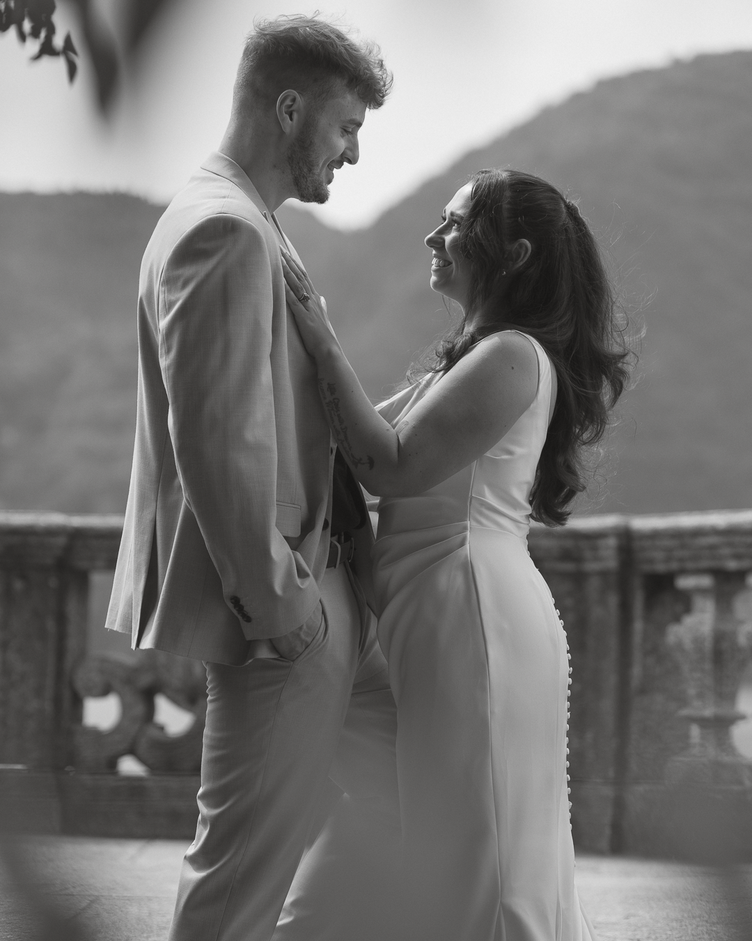 Una coppia felice si guarda innamorata, una donna sorride e ha le mani sul petto di un uomo durante un momento romantico all'aperto, con uno sfondo di montagne.