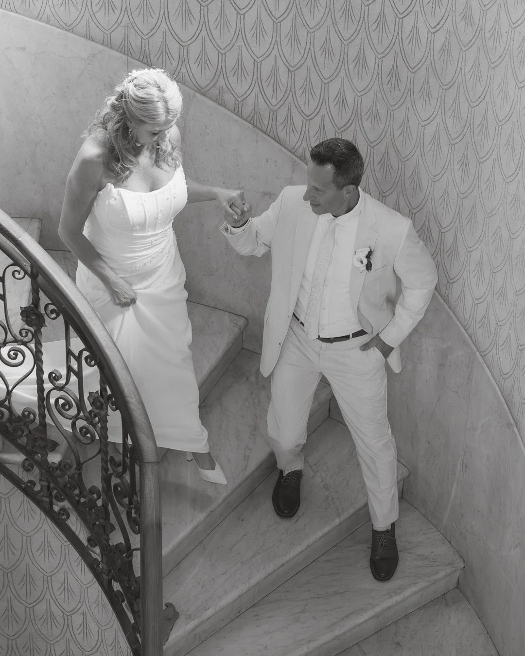 Una coppia in abiti da matrimonio, una donna in un vestito bianco e un uomo in completo bianco, si tengono per mano mentre salgono una scala di marmo con ringhiera in ferro battuto, in un ambiente elegante.