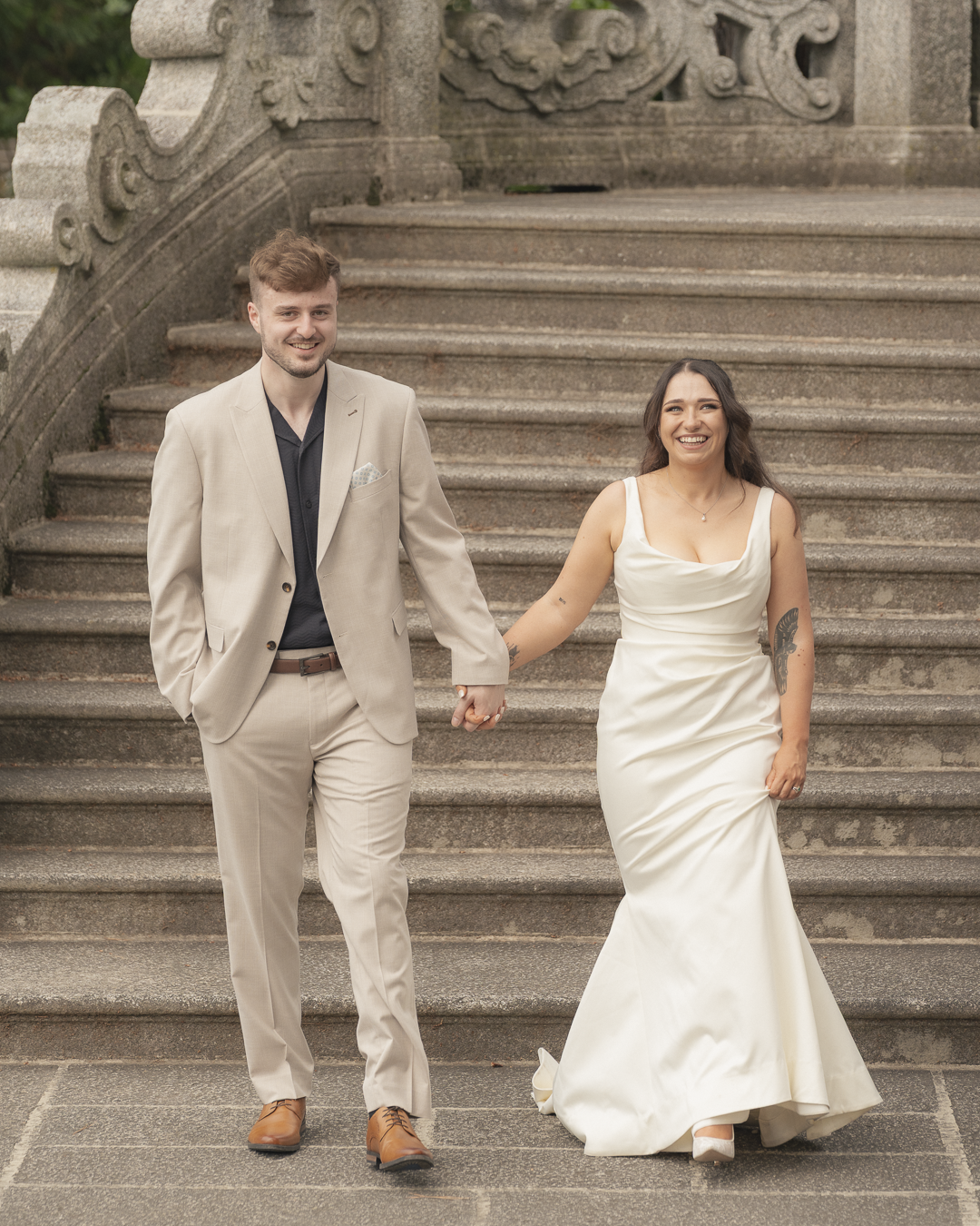 Una coppia di sposi felice cammina mano nella mano su delle scale di pietra in un ambiente esterno. Lui indossa un completo beige e lei indossa un abito da sposa bianco.
