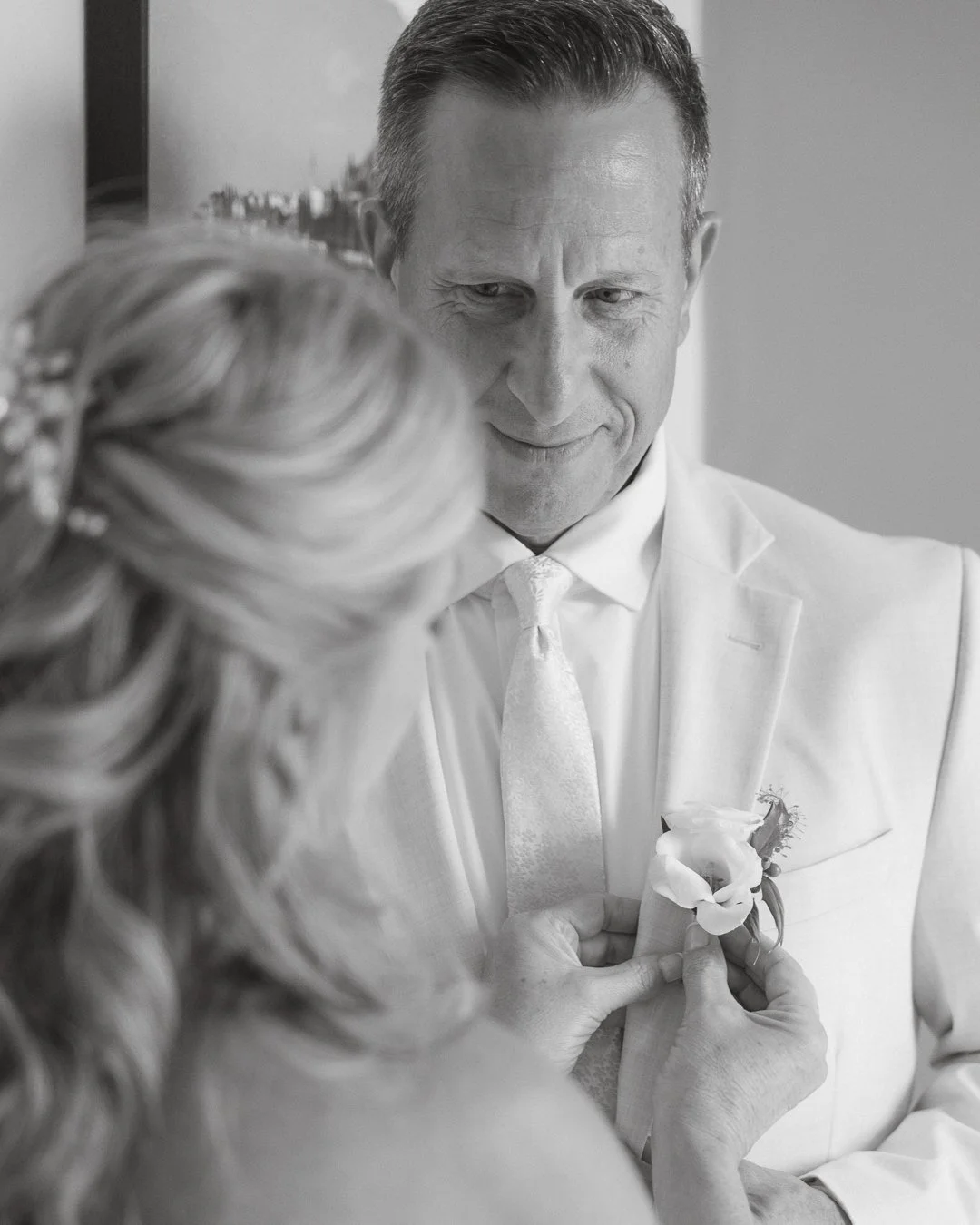 Un uomo e una donna, sembra un matrimonio, si sorridono mentre la donna si sta facendo cucire una rosa sulla giacca dell'uomo. La foto è in bianco e nero.