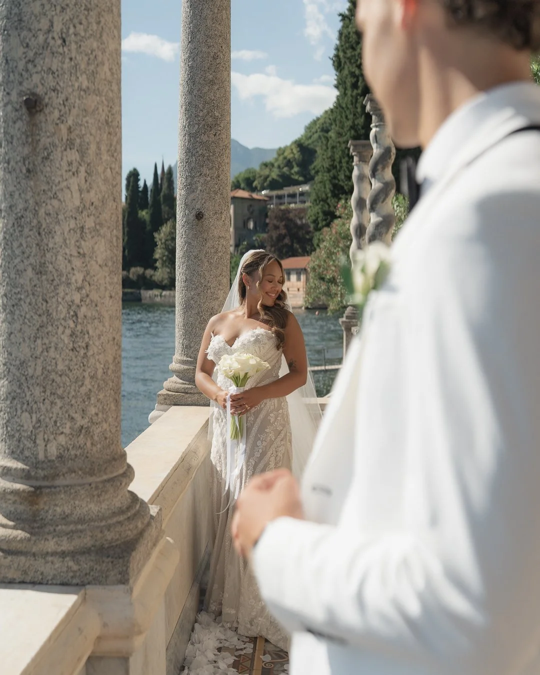 Una coppia di sposi in matrimonio, con la sposa che sorride e tiene un mazzo di fiori bianchi, mentre l'uomo, vestito di bianco, si trova più vicino alla fotocamera e guarda verso di lei. Sullo sfondo si vede un fiume, alberi e un paesaggio collinare