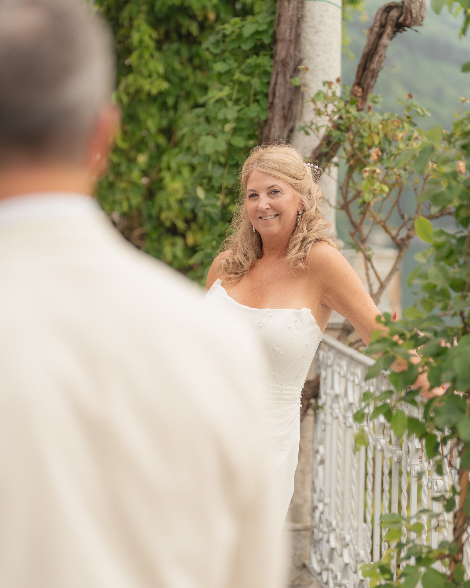 Una donna sorridente indossa un abito bianco da sposa e si trova su un balcone decorato con piante verdi, con uno sfondo di vetri e tranquilli luoghi circostanti.
