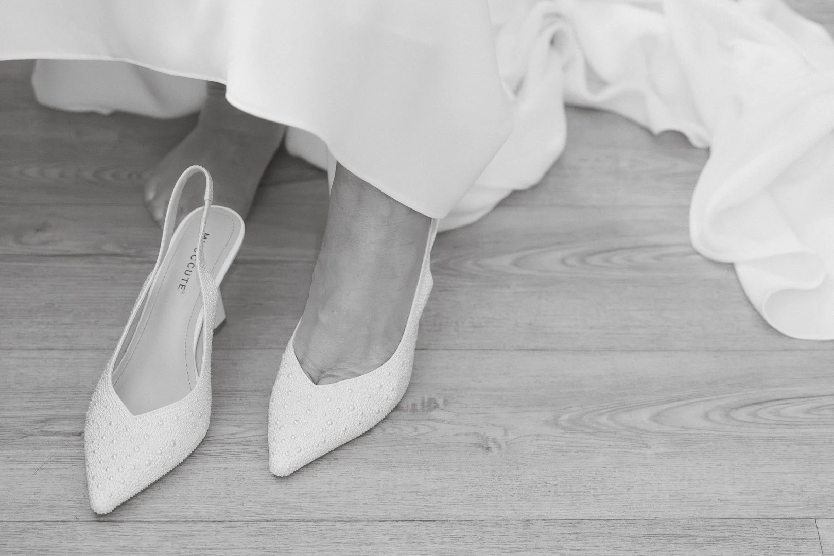 Scarpe da sposa bianche con decorazioni di perline, una delle scarpe indossata mentre l'altra è sul pavimento, una persona indossa il vestito bianco e si trova su un pavimento di legno.