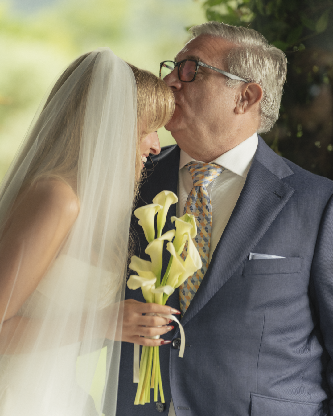 Una sposa con velo riceve un bacio sulla fronte da un uomo anziano in giacca blu durante un matrimonio.