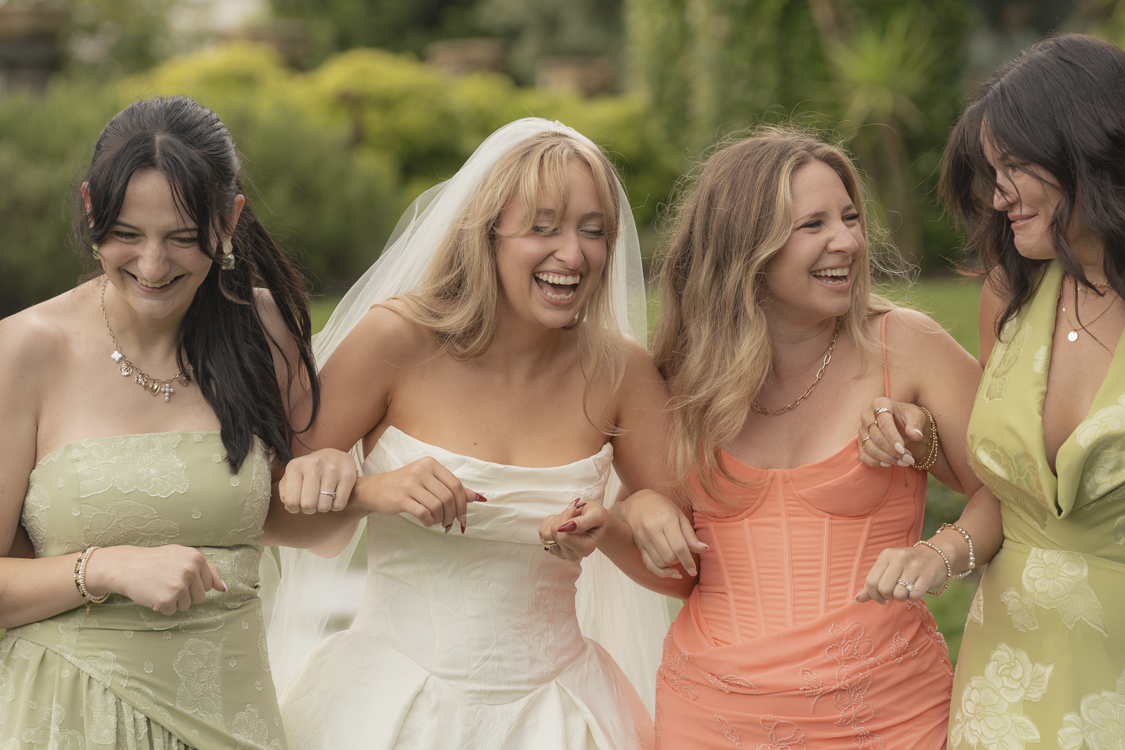 Gruppo di donne sorridenti, una in abito da sposa, che si tiene per mano all'aperto, con uno sfondo di vegetazione verde.