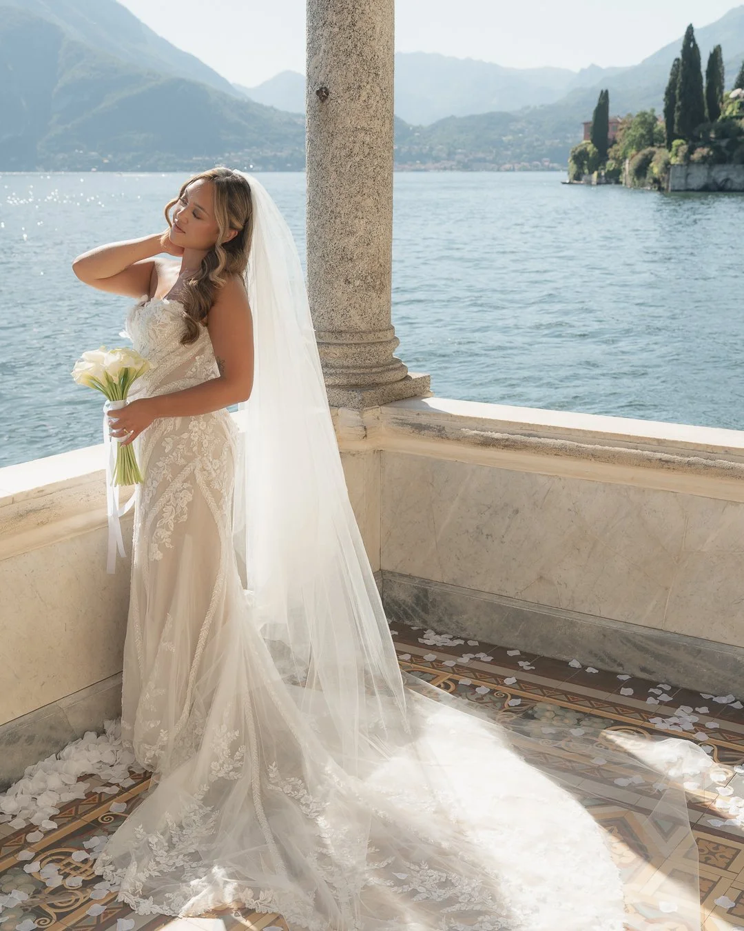 Una sposa in abito da matrimonio bianco con velo, che tiene un mazzo di calla, si trova su una terrazza con vista su un lago e montagne sullo sfondo.