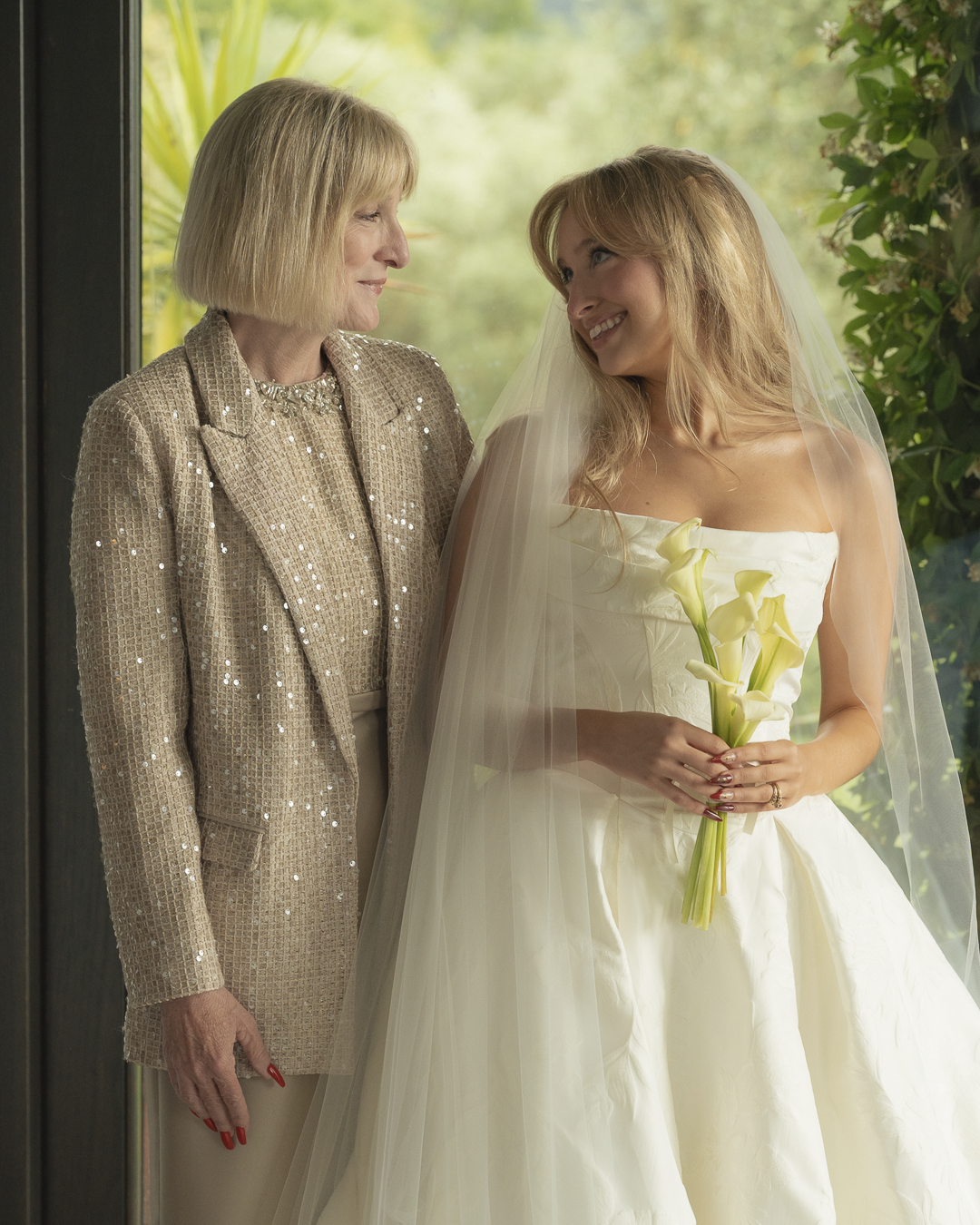 Una sposa e una donna anziana sorridenti si guardano affettuosamente, con la sposa che tiene un mazzo di calle bianche, all'interno vicino a una finestra con uno sfondo di natura verde.