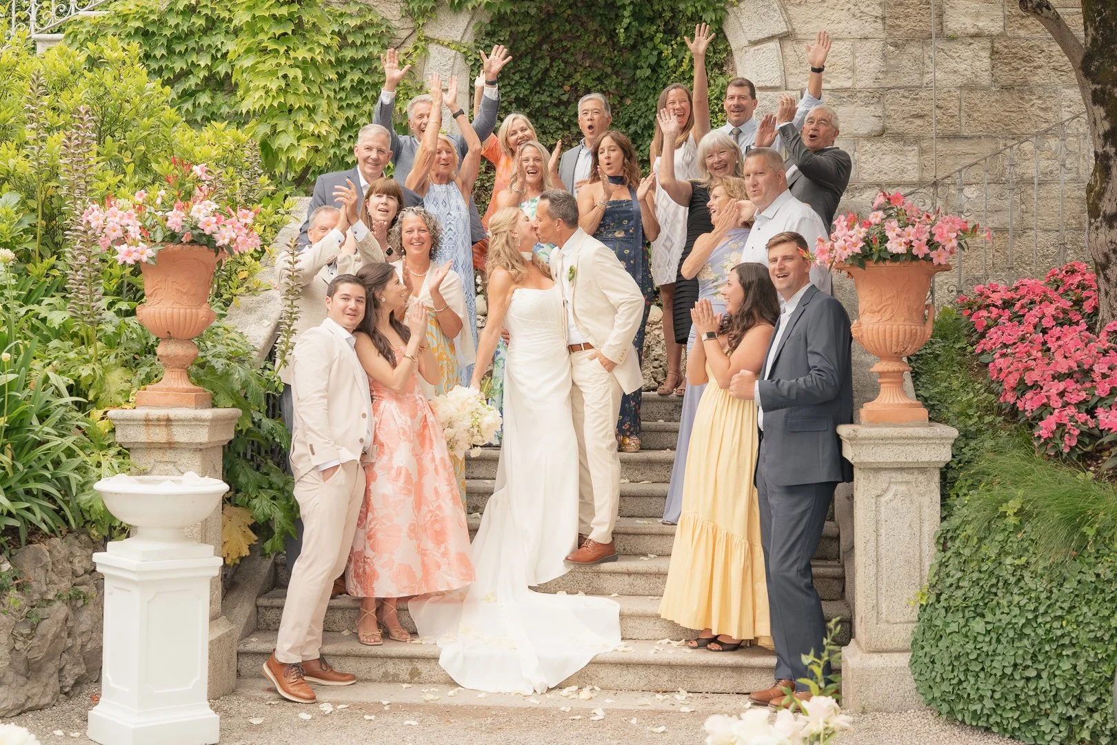 Gruppo di persone a un matrimonio sulla scalinata di un giardino, alcune scambiano baci, altri festeggiano con le mani alzate, decorazioni floreali. La coppia di sposi al centro si bacia.
