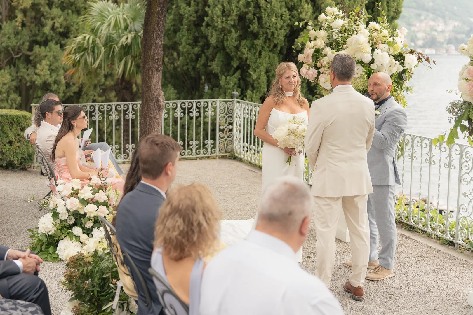 Coppia di sposi che si scambia le promesse di matrimonio durante una cerimonia all'aperto con fiori e vista sul lago, con amici e parenti seduti che assistono all'evento.