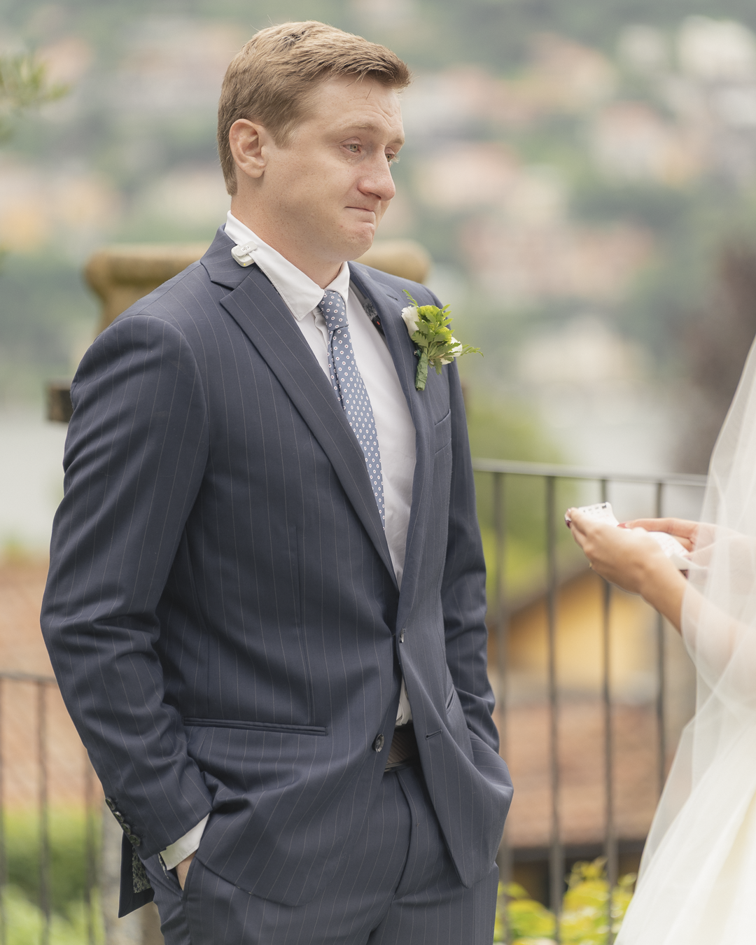 Un uomo vestito con abito da cerimonia blu scuro con strisce sottili, con una camicia bianca e una cravatta blu con pois bianchi, durante un matrimonio. È emozionato e triste, probabilmente mentre aspetta di essere amato dalla sposa, che non è visibl