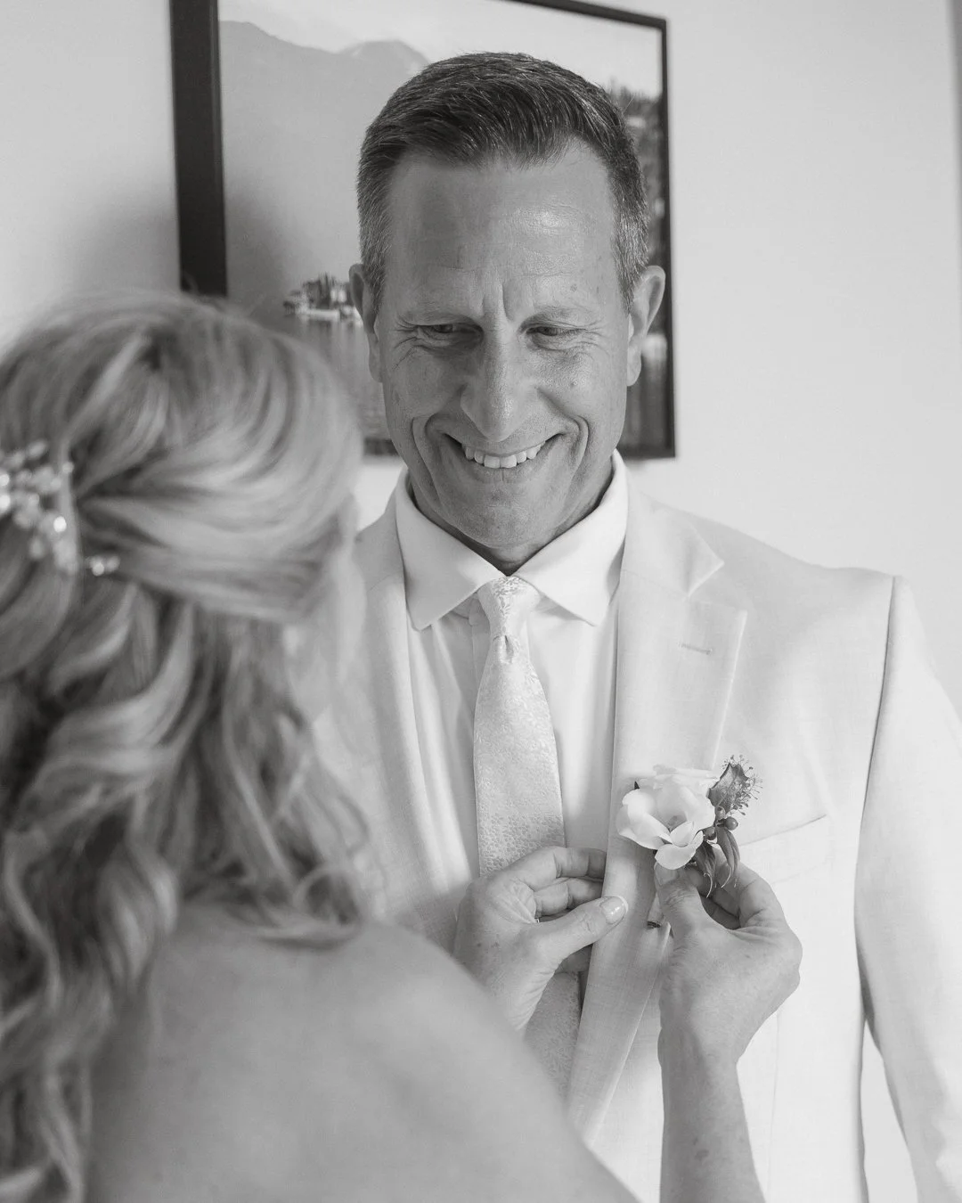 Un uomo sorridente indossa un abito chiaro e una cravatta, una donna è mentre gli attacca una boutonnière sul petto, presumibilmente al matrimonio.