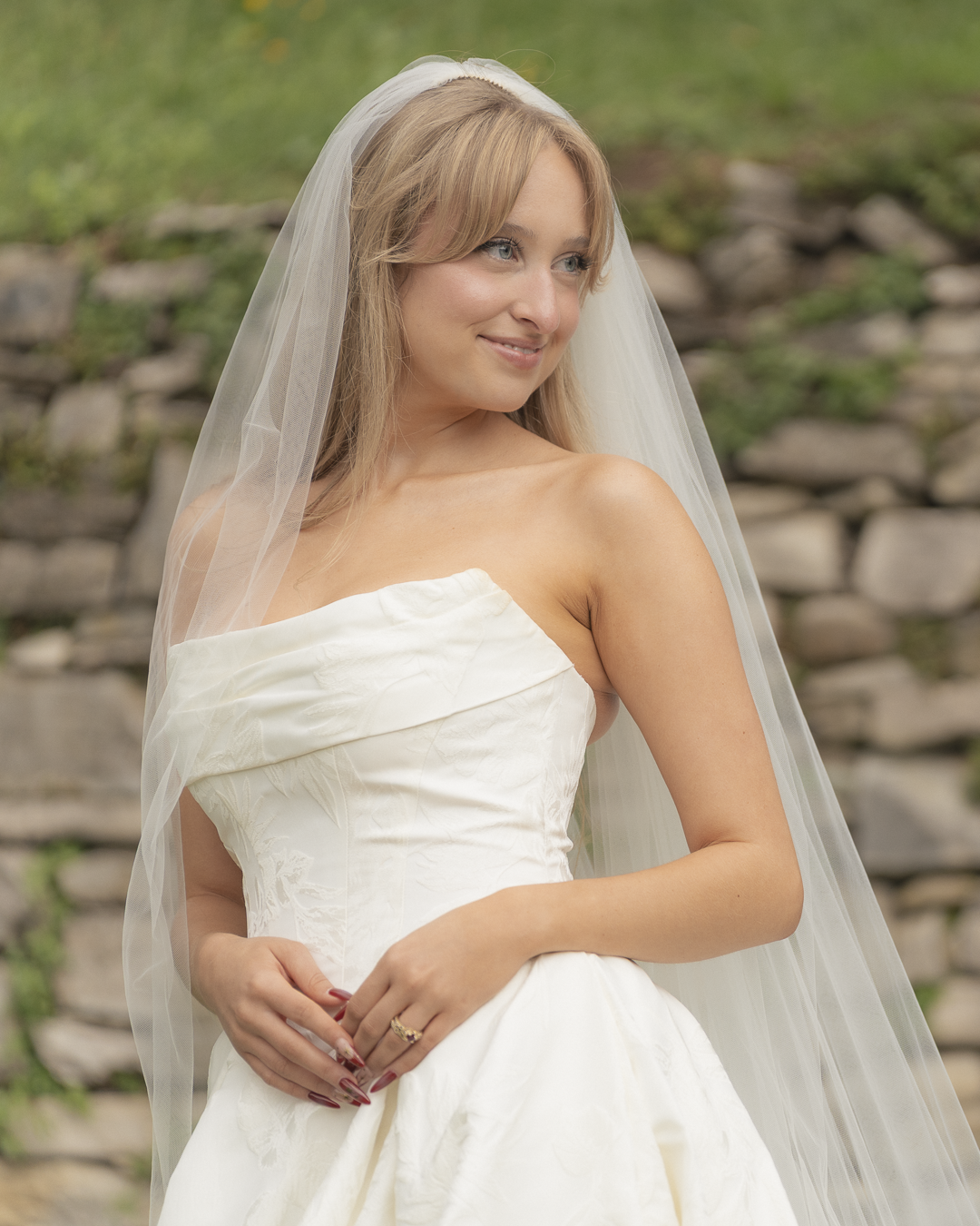 Una donna in abito da sposa con velo bianco, che sorride e guarda di lato, con uno sfondo di pietre e vegetazione.