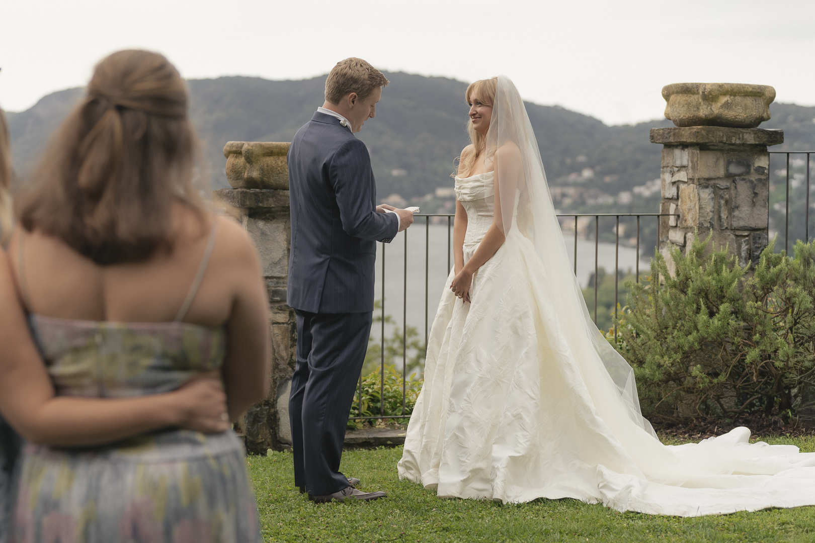 Spose di matrimonio con il testimone che legge una promessa, sposa in abito bianco e vedovo in abito scuro, sullo sfondo un paesaggio di colline e acqua.