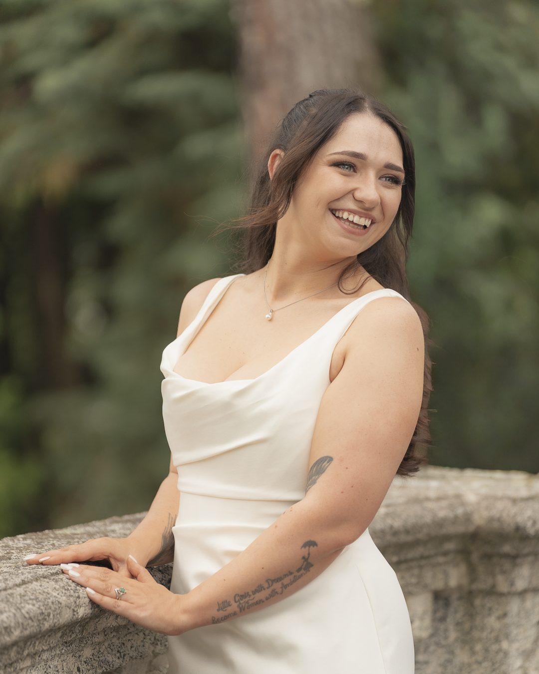 Una donna sorridente con capelli castani e tatuaggi su braccia, indossa un vestito bianco e una collana, si trova in un ambiente naturale con alberi sullo sfondo.