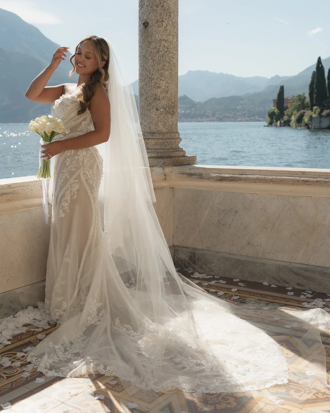 Sposa sorridente con abito da matrimonio, veffuto di fiori bianchi, in piedi su una balconata con vista lago e montagne in background.