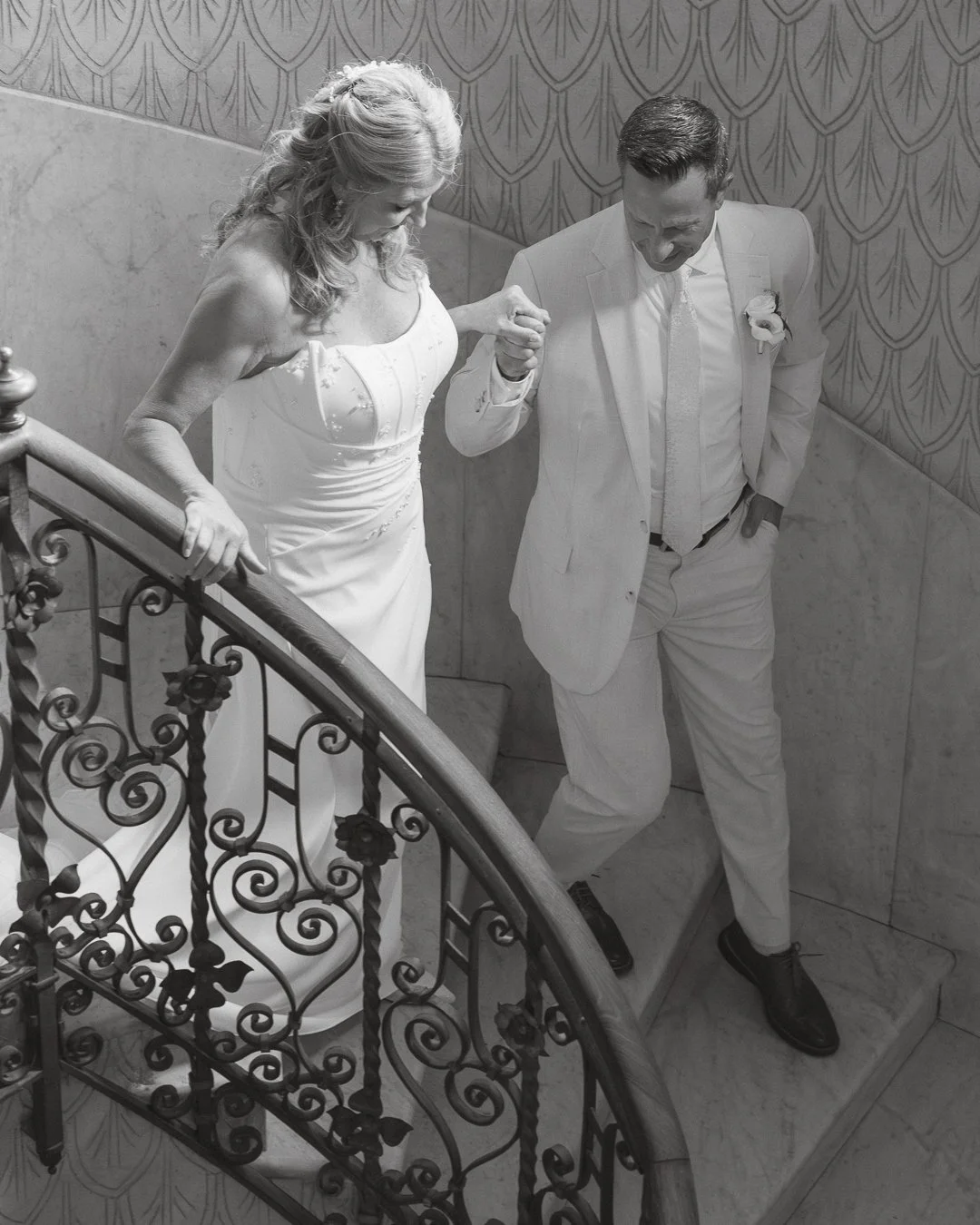 Una coppia di persone in abiti da matrimonio, una donna in abito bianco e un uomo in completo bianco, che camminano insieme lungo una scala a spirale con ringhiera decorativa, in un ambiente interno.