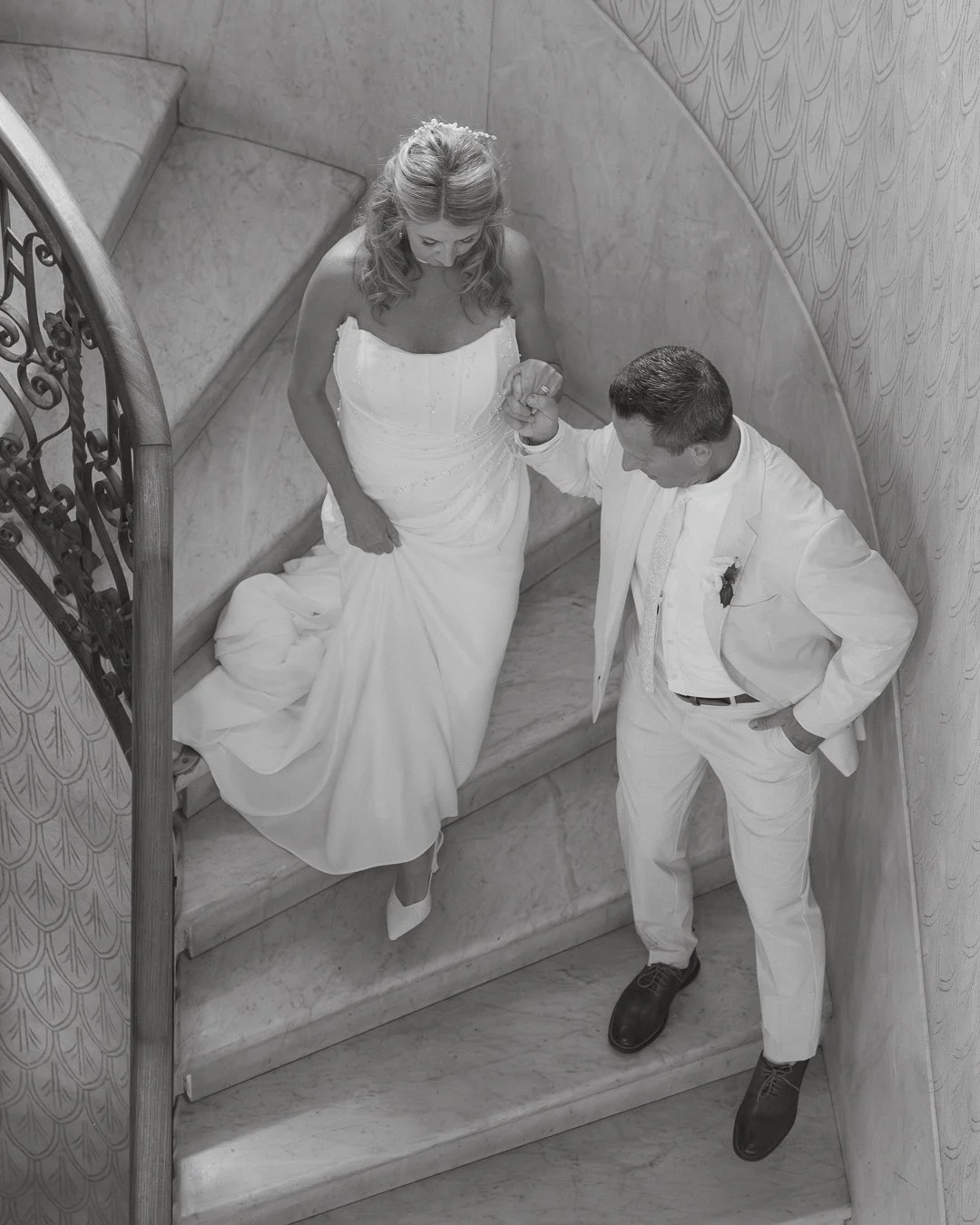 Una coppia in abiti nuziali si abbraccia sulla scala di marmo con una donna in un vestito bianco e un uomo in completo bianco, in un ambiente elegante interiore.