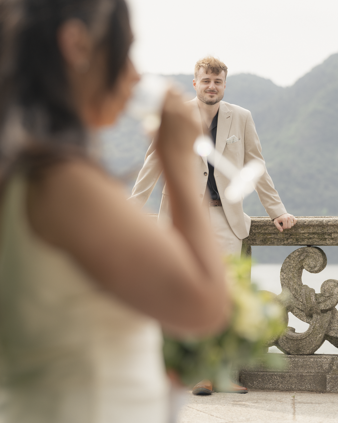 Un uomo in giacca beige sorride guardando una donna in primo piano che tiene un fiore, sullo sfondo un paesaggio di montagna.