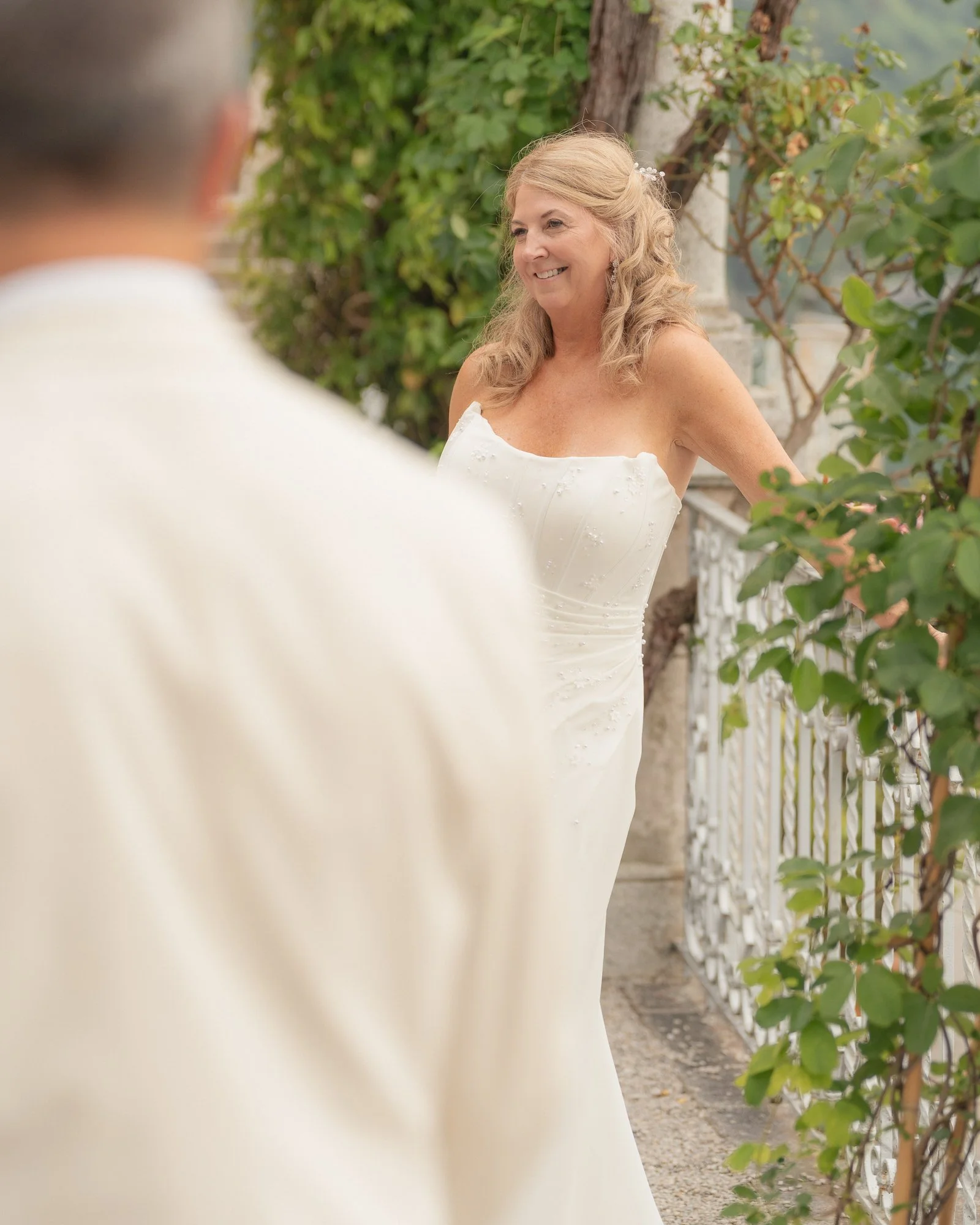Una donna sorridente indossa un abito bianco da sposa, seduta vicino a un cancello decorativo in un giardino con piante verdi.