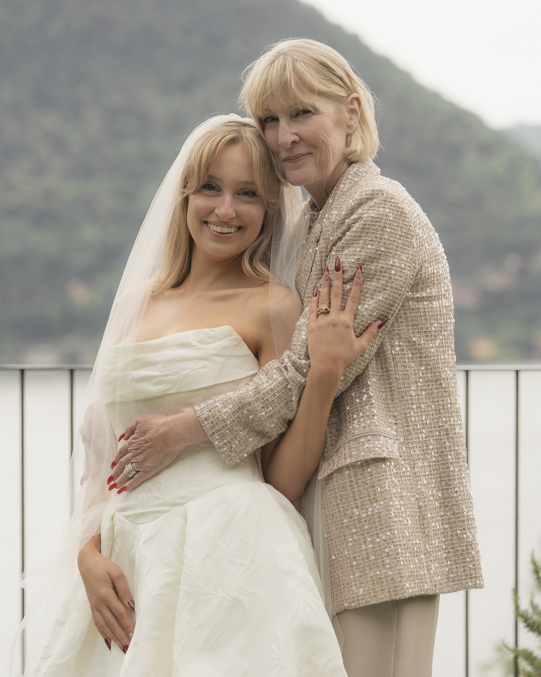 Una sposa in abito bianco e un'anziana signora sorridenti si abbracciano davanti a un mare con montagne sfocate sullo sfondo.