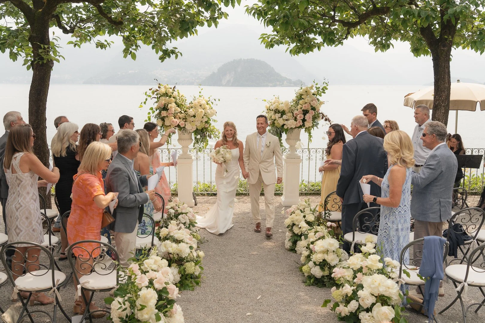 Coppia di sposi che si tiene per mano durante il rito di matrimonio all'aperto, circondati da amici e parenti, con un lago e delle montagne sullo sfondo, decorazioni floreali e un grande vaso di fiori davanti all'altare.