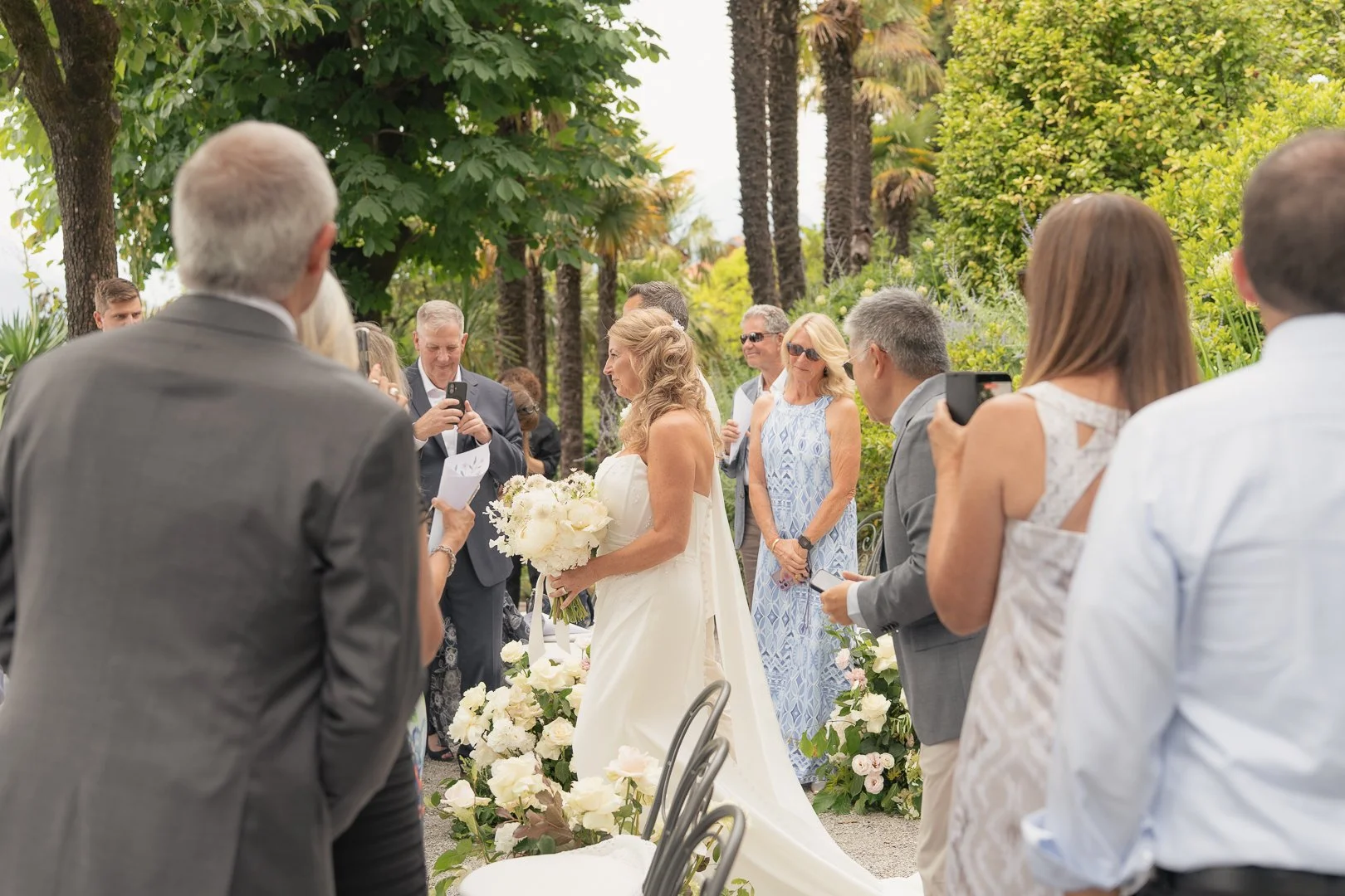 Una coppia di sposi durante il loro matrimonio all'aperto, circondati da amici e parenti, con molte persone che scattano foto o registrano il momento speciale, in un giardino con alberi e piante verdi.