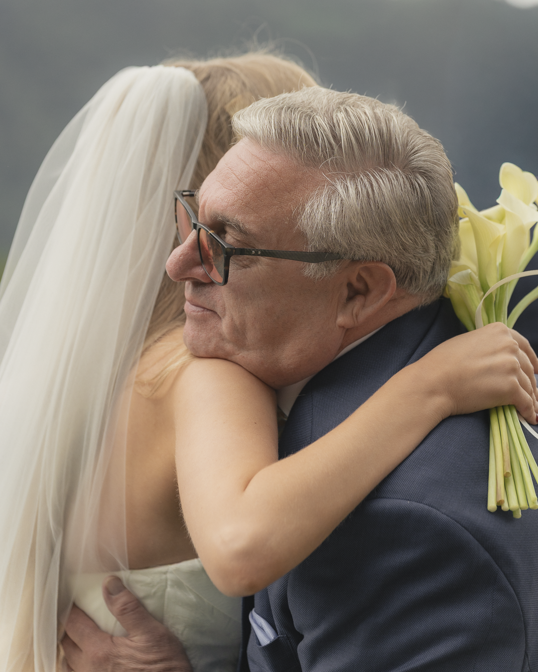 Un anziano uomo e una donna, presumibilmente sposi, si abbracciano strettamente in un momento emotivo, con quest'ultima che tiene un mazzo di calle bianche.
