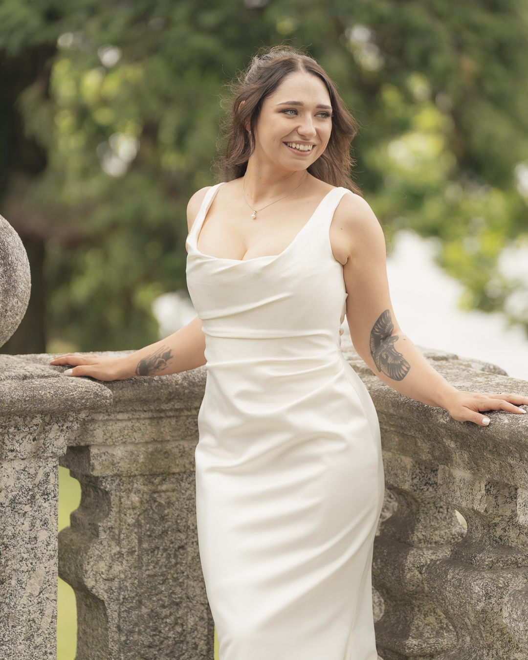 Una donna sorridente indossa un abito da sposa bianco, si appoggia a una balaustra di pietra in un ambiente esterno con alberi verdi sullo sfondo.