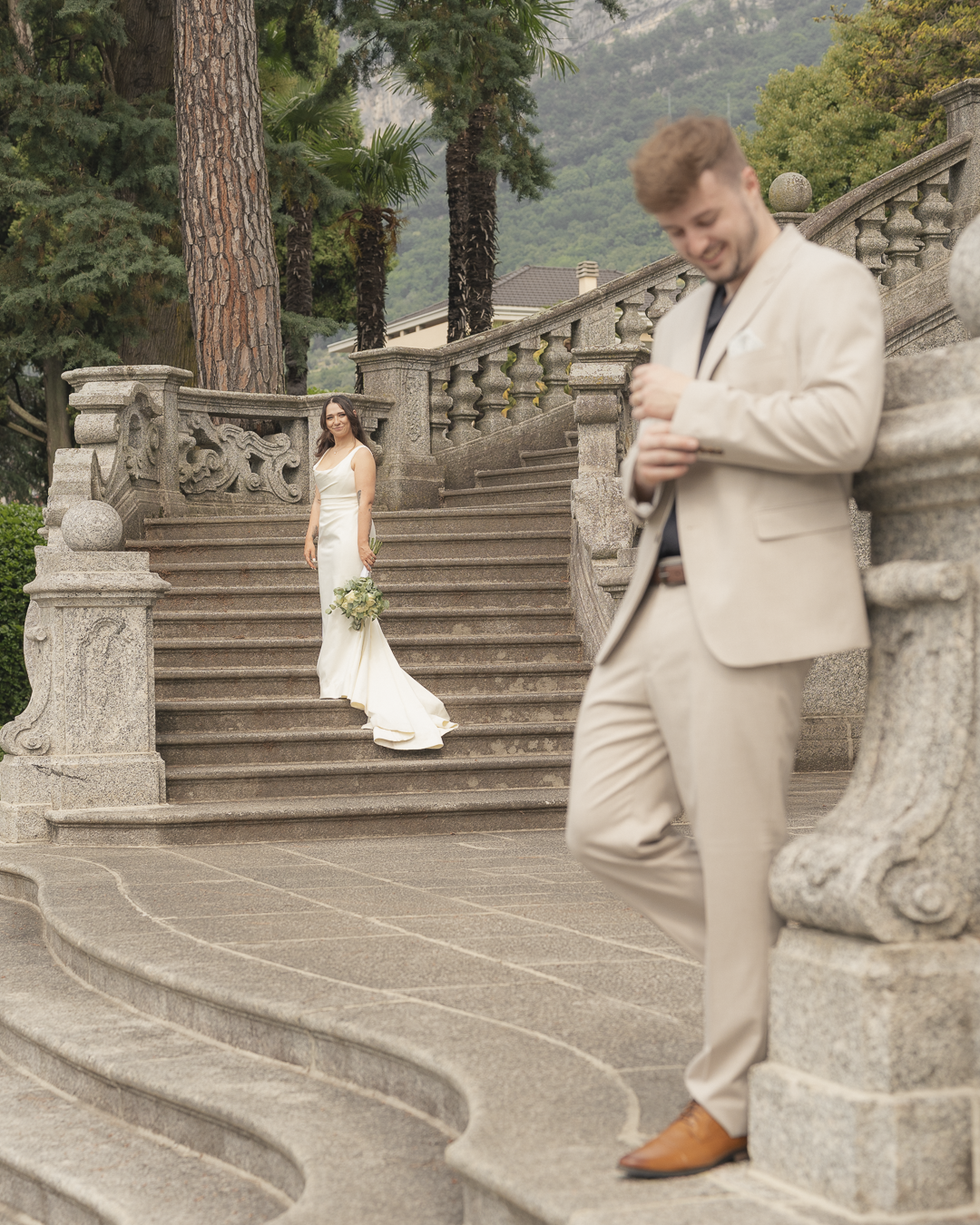 Una coppia di sposi in un matrimonio all'aperto, con la sposa in piedi sulla scala tenendo un bouquet e l'uomo sorridente che si appoggia a un murello, sotto alberi e un paesaggio montano.