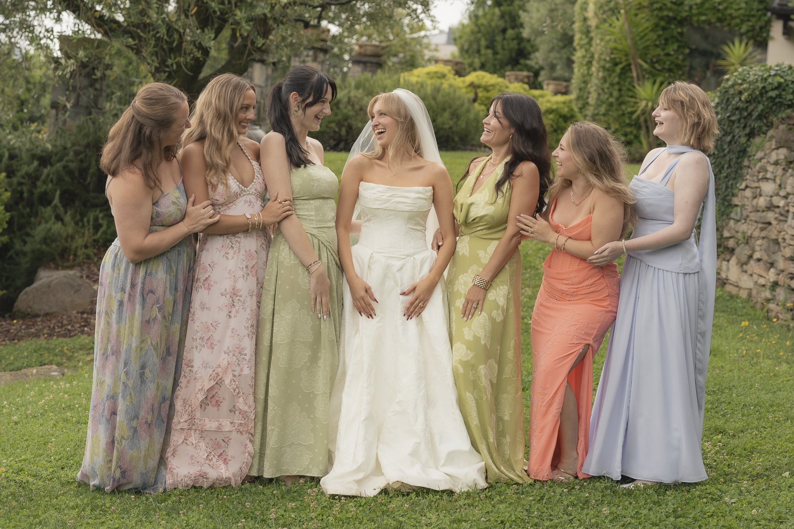 Gruppo di donne e una sposa sorridenti in un giardino verde, durante una cerimonia di matrimonio, abiti eleganti e colorati.