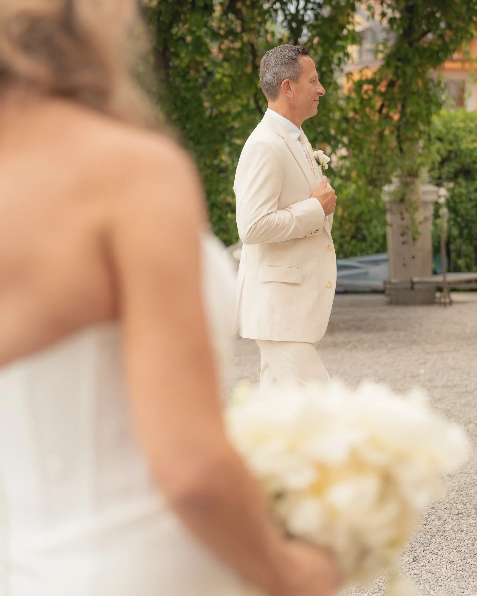 Un uomo indossa un abito beige, probabilmente durante un matrimonio, e sta davanti a uno sfondo di alberi e edifici, in un ambiente all'aperto.