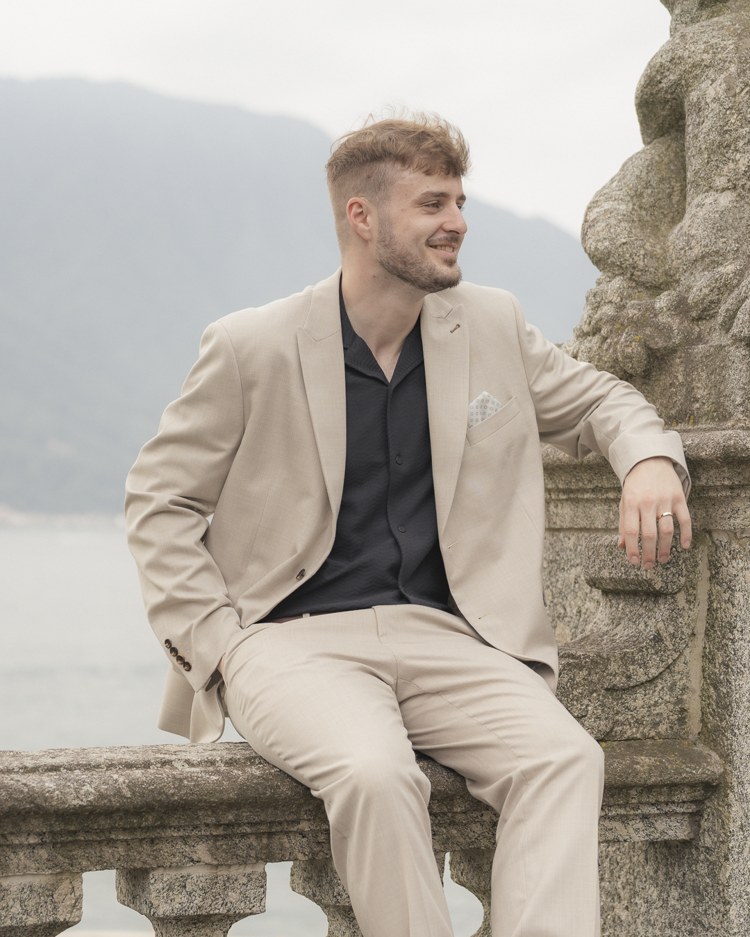 Un uomo seduto su una ringhiera di pietra, indossa un completo beige e una camicia nera, con uno sfondo di montagne e acqua