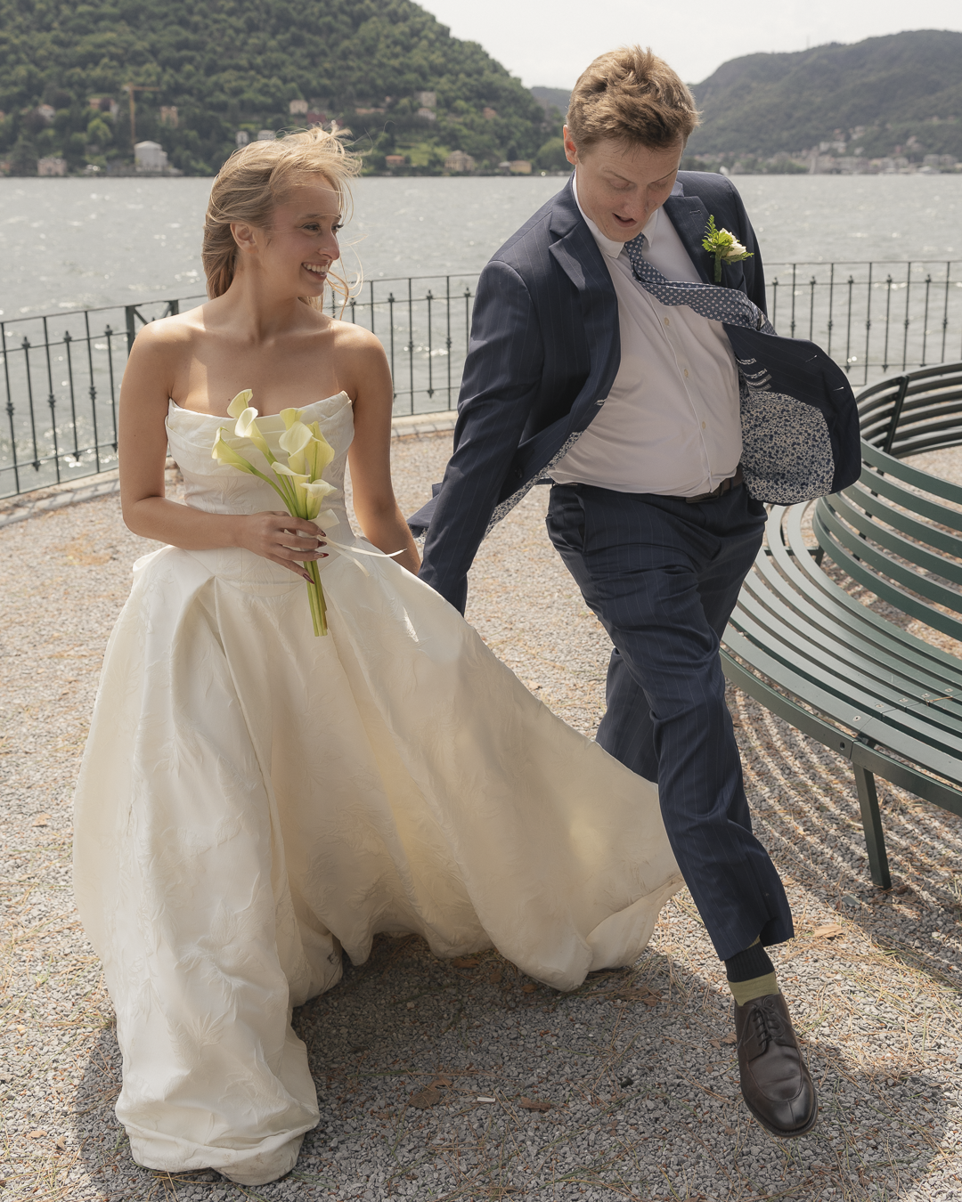 Una coppia felice di sposi arrampicata su una panchina vicino a un lago con montagne sullo sfondo, durante il loro matrimonio.
