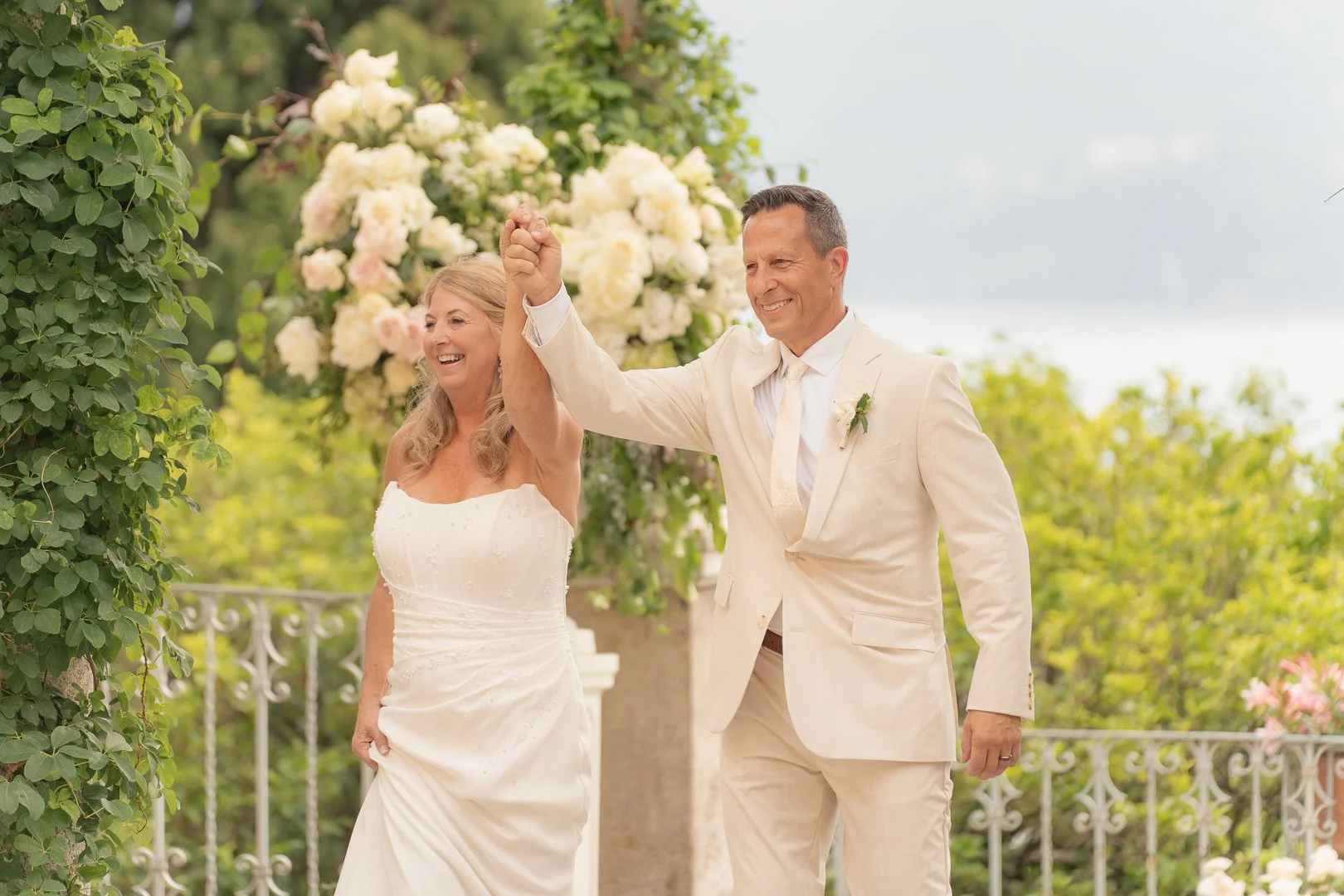 Una coppia di sposi felici durante il loro matrimonio all'aperto, con pannocchie di fiori bianchi sullo sfondo, che si tengono per mano sorridendo.
