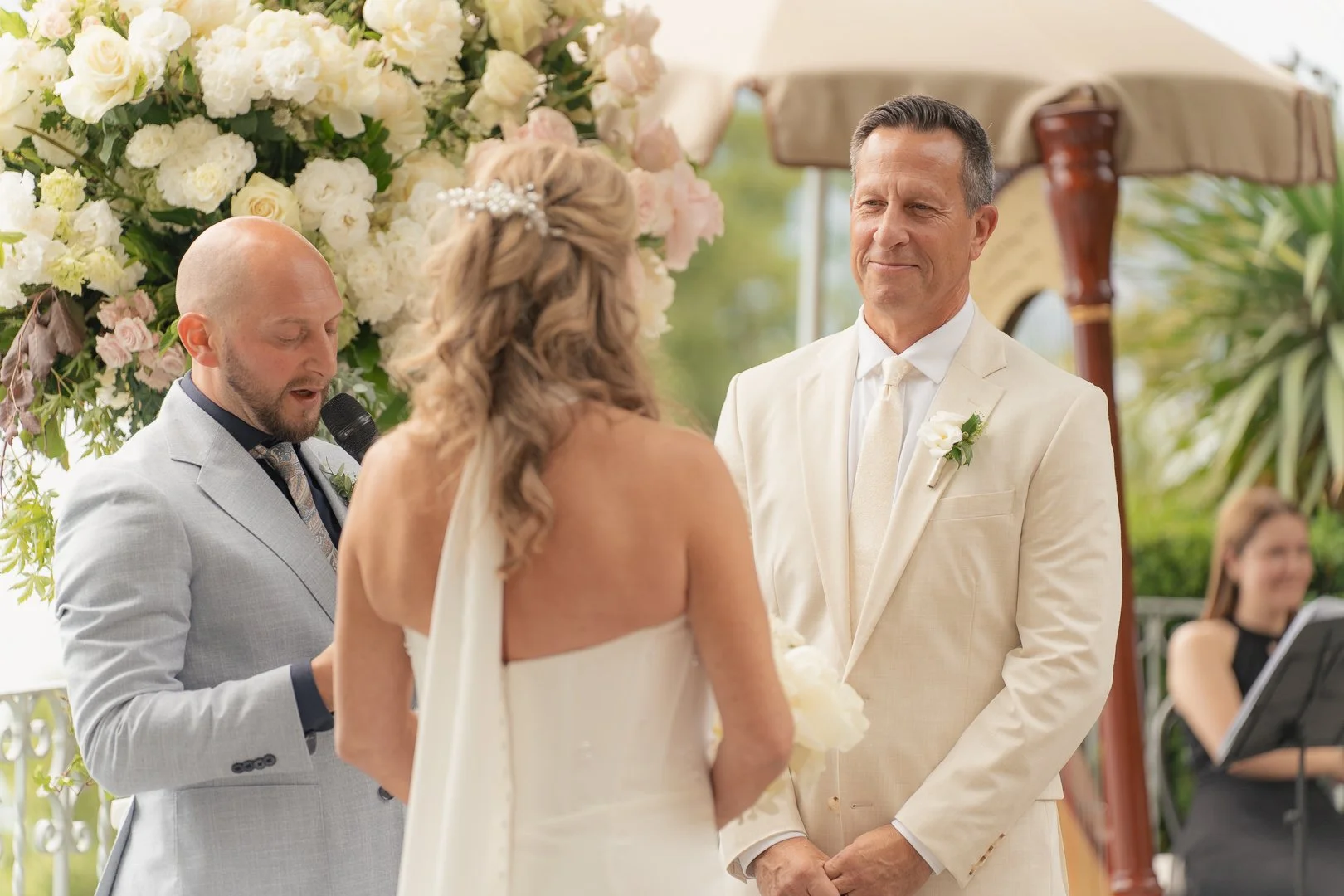Una coppia di sposi durante il matrimonio, con l'uomo in abito bianco e la donna in abito da sposa, si scambiano i voti davanti all'ufficiale di matrimonio, con decorazioni floreali e un pubblico di invitati sullo sfondo.
