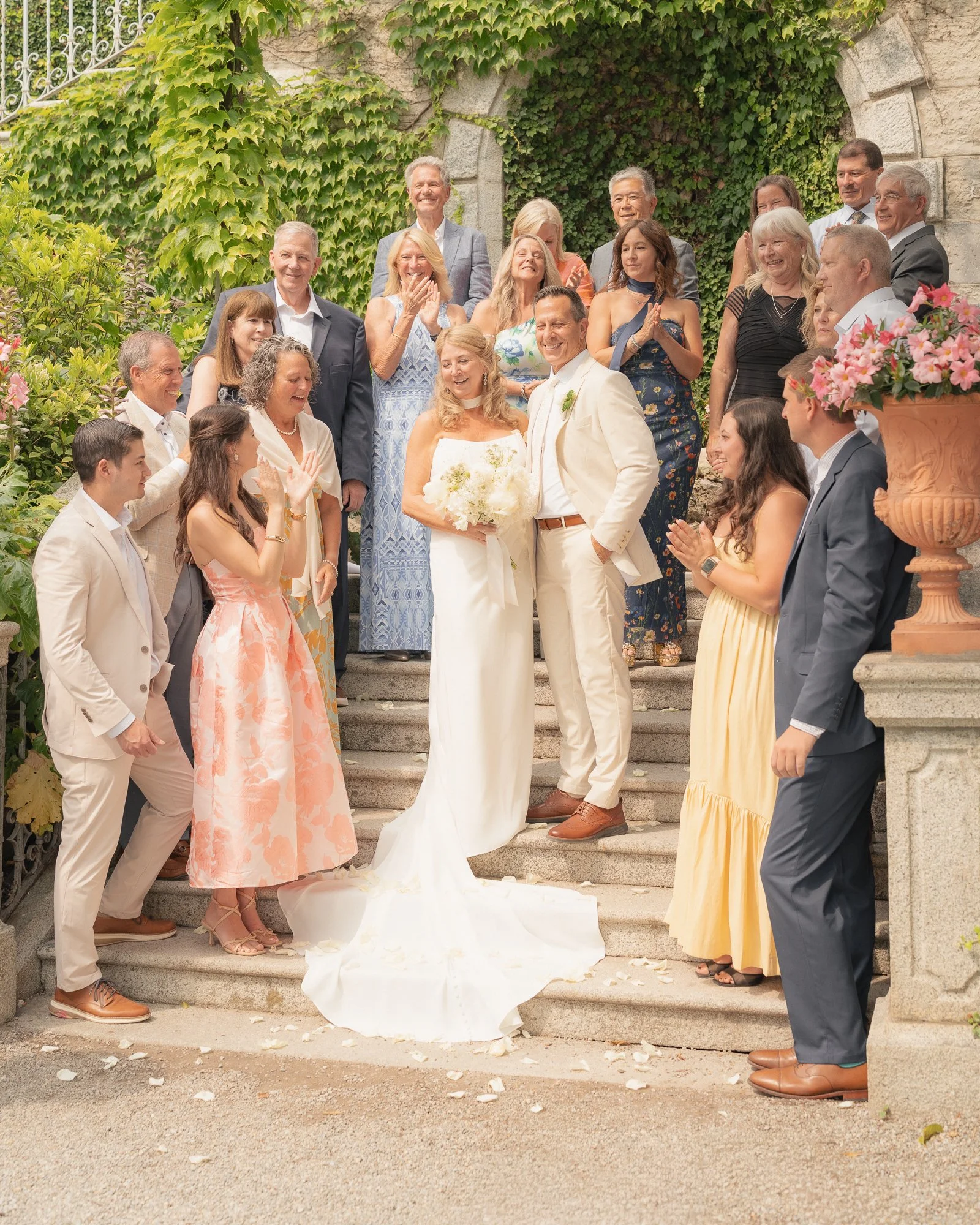 Un gruppo di persone sorridenti e felici sono riunite per un matrimonio sulla scalinata di un edificio storico con pareti in pietra e muri ricoperti di edera verde. Al centro, una coppia di sposi con abiti eleganti, circondati da amici e familiari. S