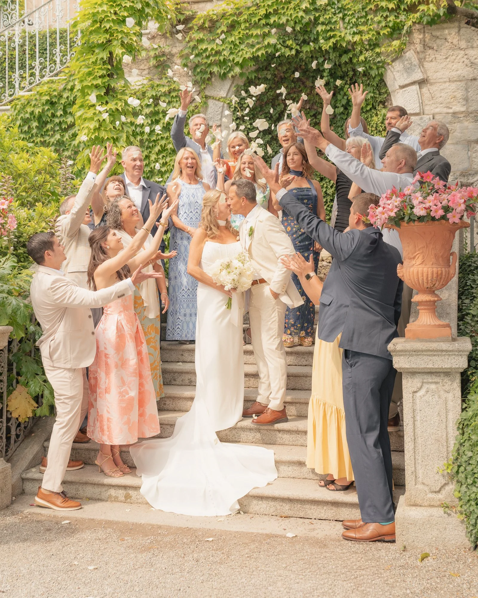 Gruppo di persone ai festeggiamenti di un matrimonio, con alcuni che spruzzano petali e altri che si baciano sulla bocca, immersi tra vegetazione e fiori